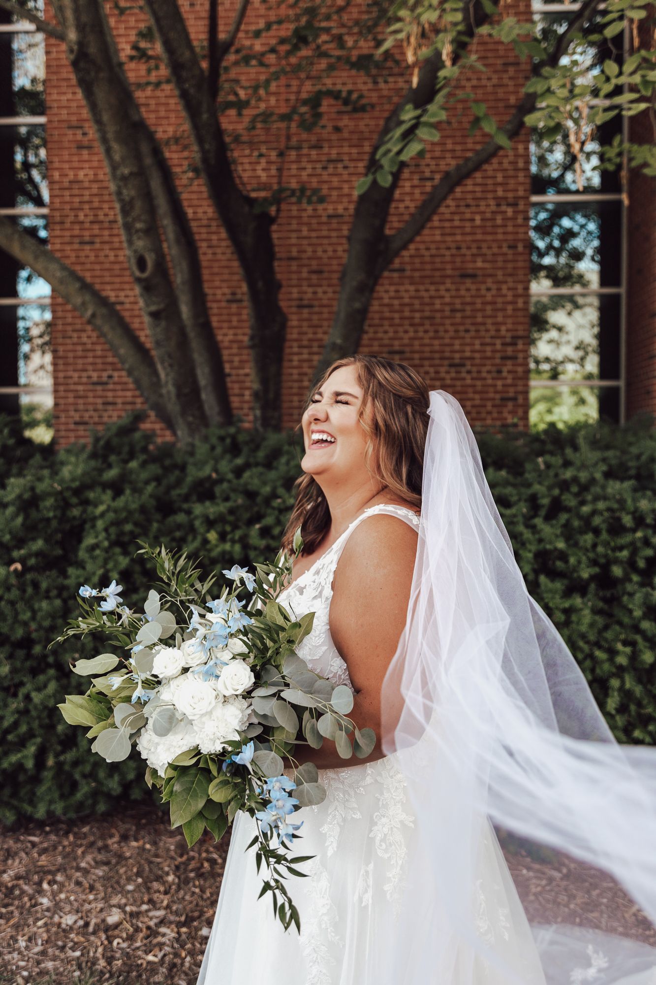 Joyful bride laughs while showing off her bouquet and veil against brick wall backdrop.