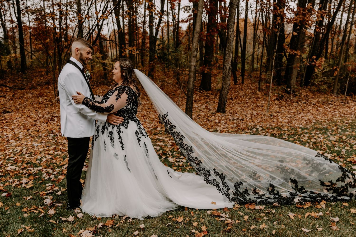 Autumn wedding portrait with bride in lace dress and dramatic long veil beside groom in white jacket.