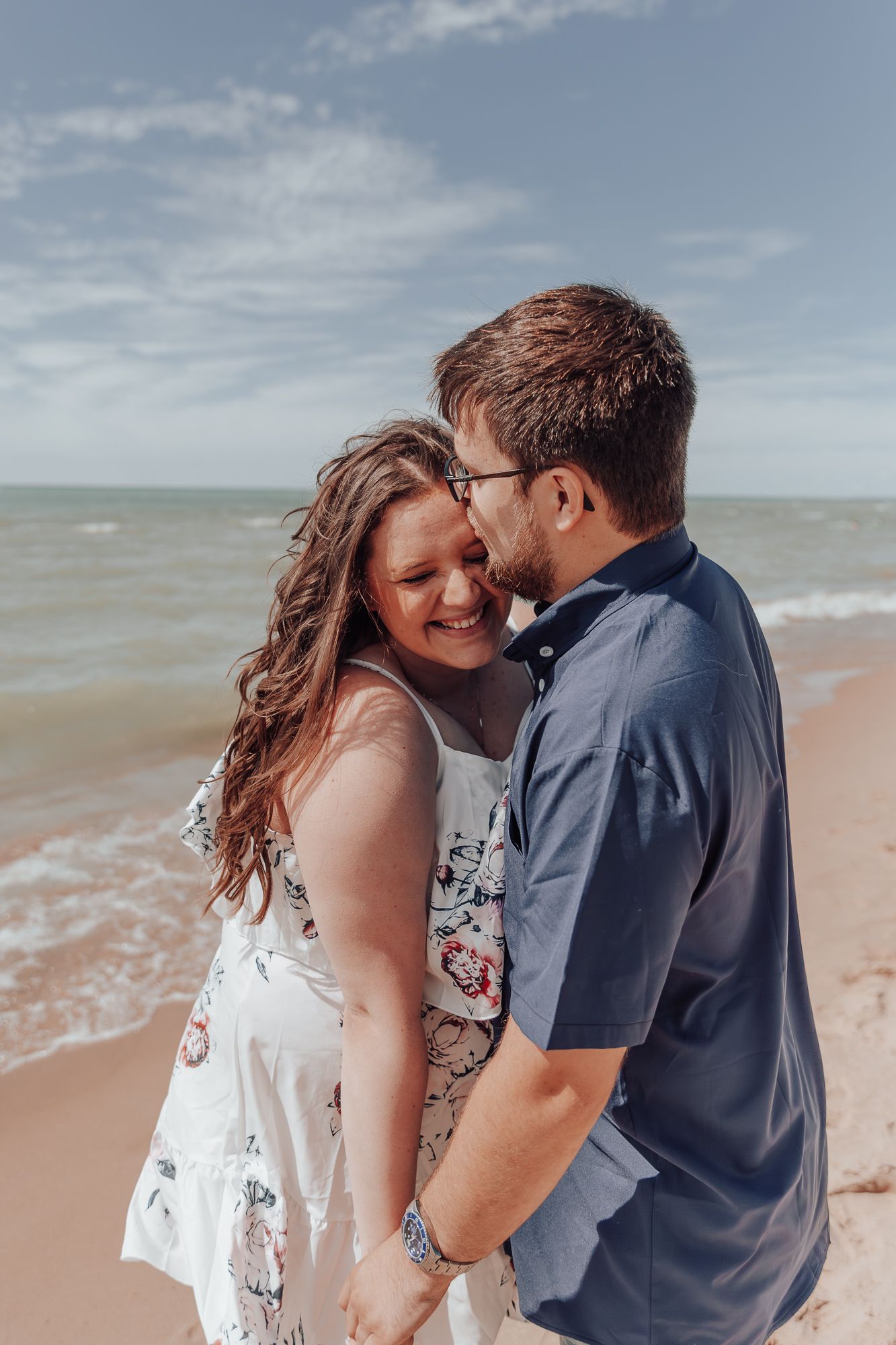 A romantic couple embraces and smiles on a sunny beach with ocean waves in the background.