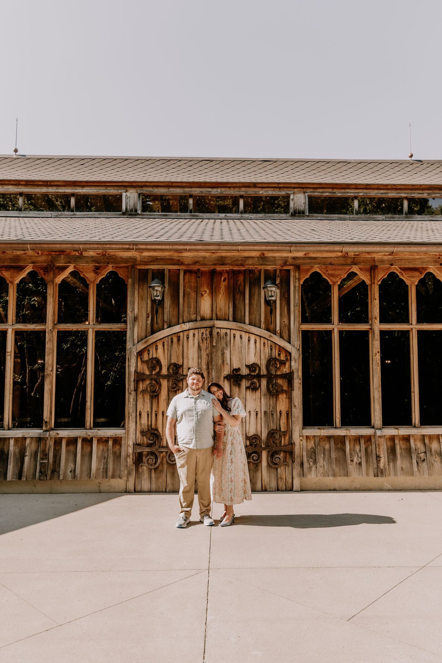 Couple posing in front of rustic wooden barn venue with arched doors on a sunny day.