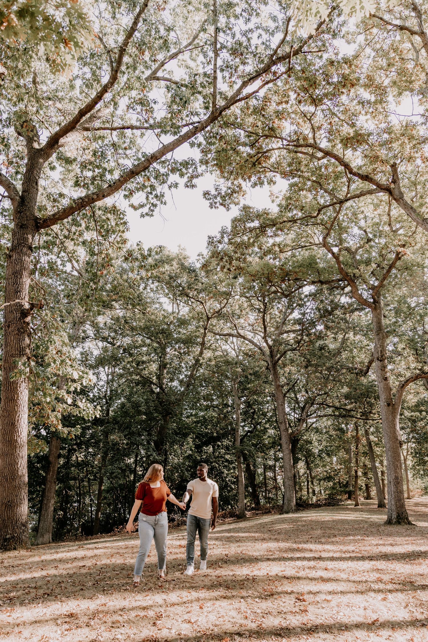 A couple walks hand in hand down a scenic tree-lined path through a peaceful autumn forest.