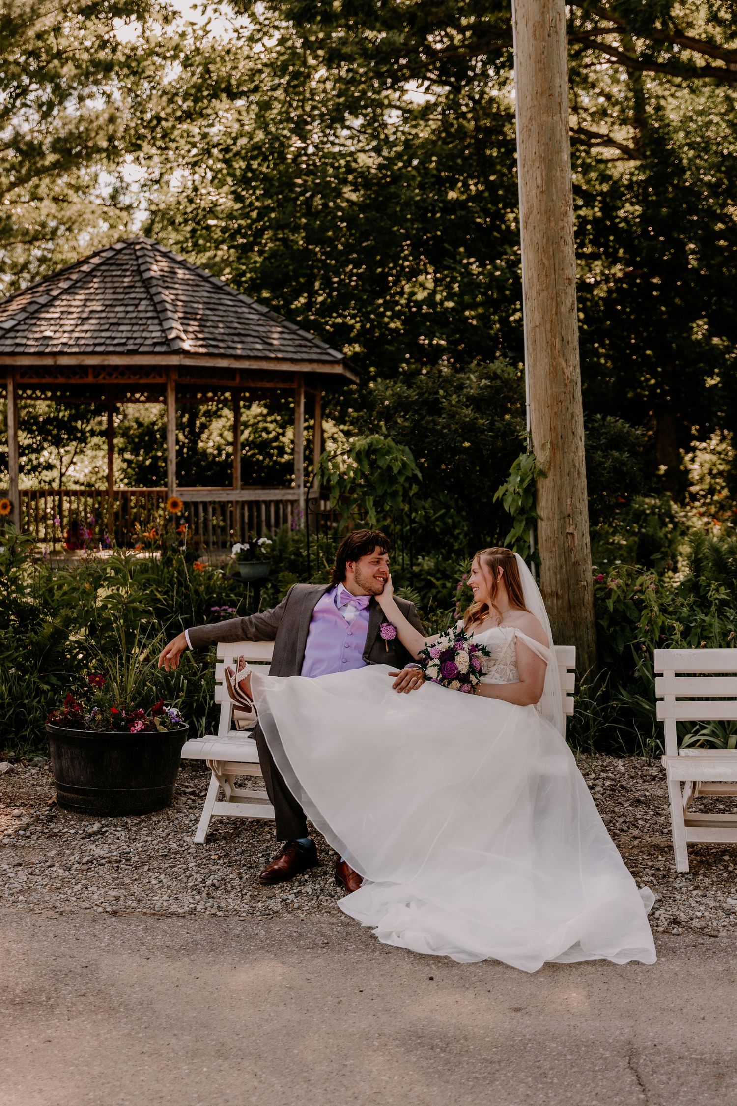 Bride and groom sitting on a white bench near a gazebo surrounded by lush greenery on their wedding day.