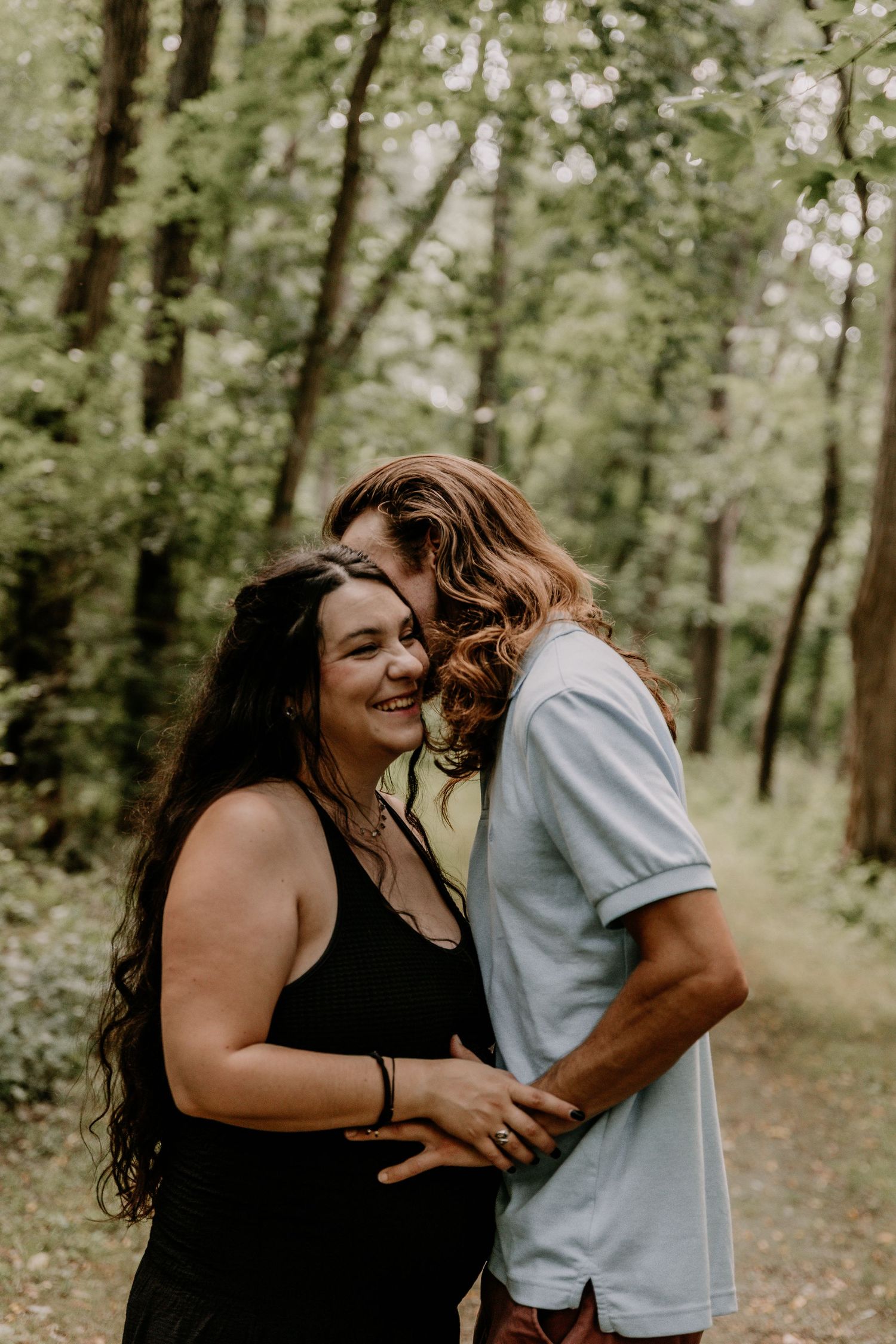 A couple embraces on a forest path, the woman in black smiling joyfully as her partner nuzzles her hair.