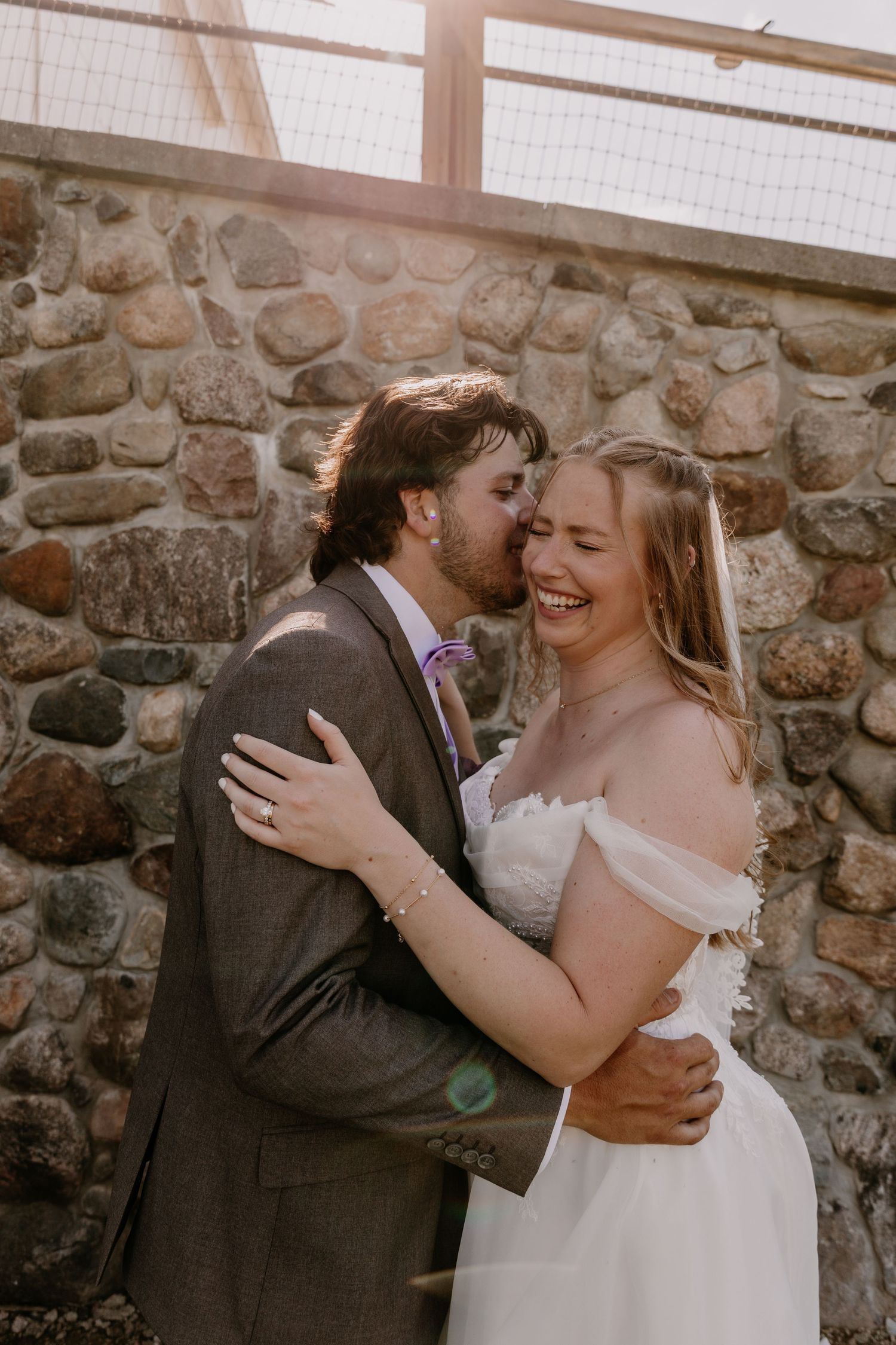 A joyful bride in an off-shoulder white gown laughs as her groom in a gray suit kisses her cheek near a stone wall.