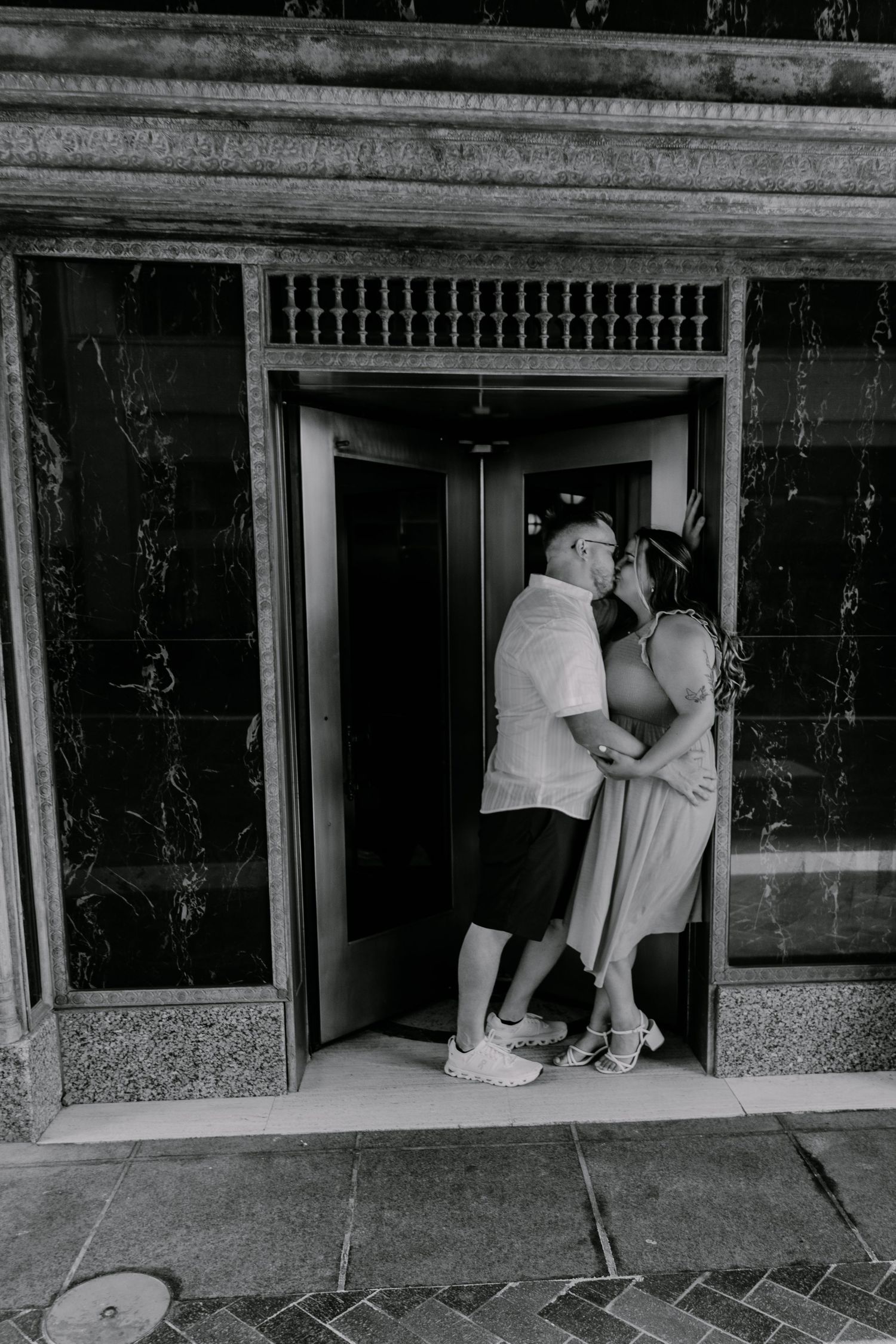 Black and white photo showing two people peering through a vintage building entrance doorway with marble walls.