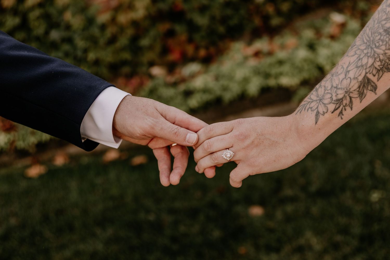 Two hands wearing wedding rings touch fingers tenderly against a blurred green garden background.