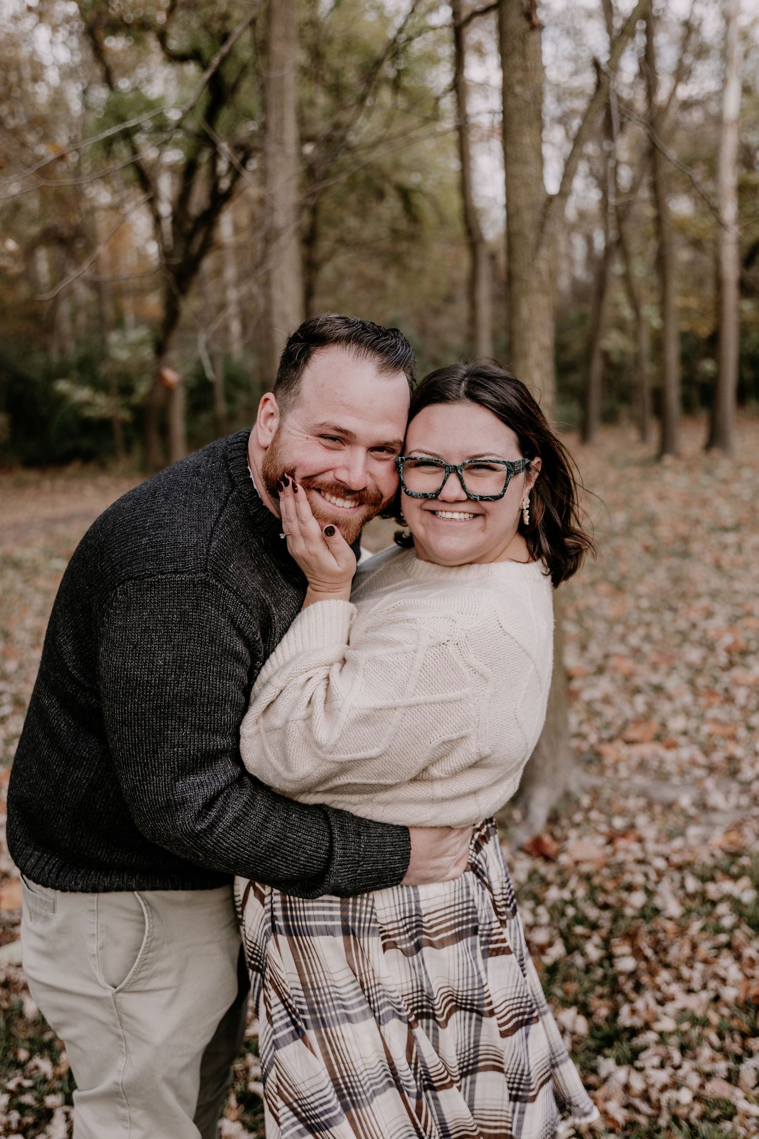 A couple shares intimate moments in a wooded autumn setting wearing cozy sweaters among fallen leaves.