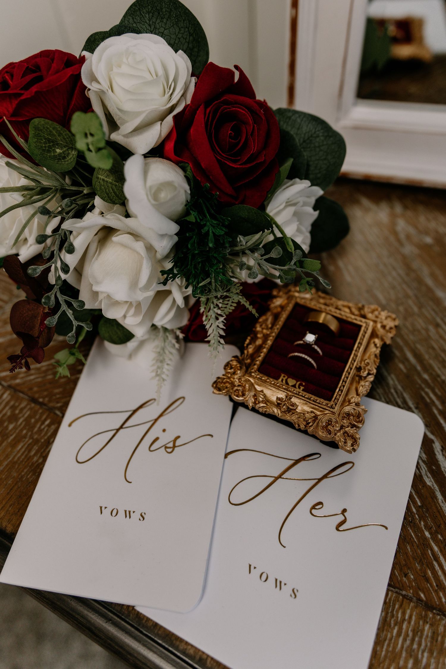 Wedding vows booklets labeled His and Hers beside a diamond ring in gold frame and red and white rose bouquet.