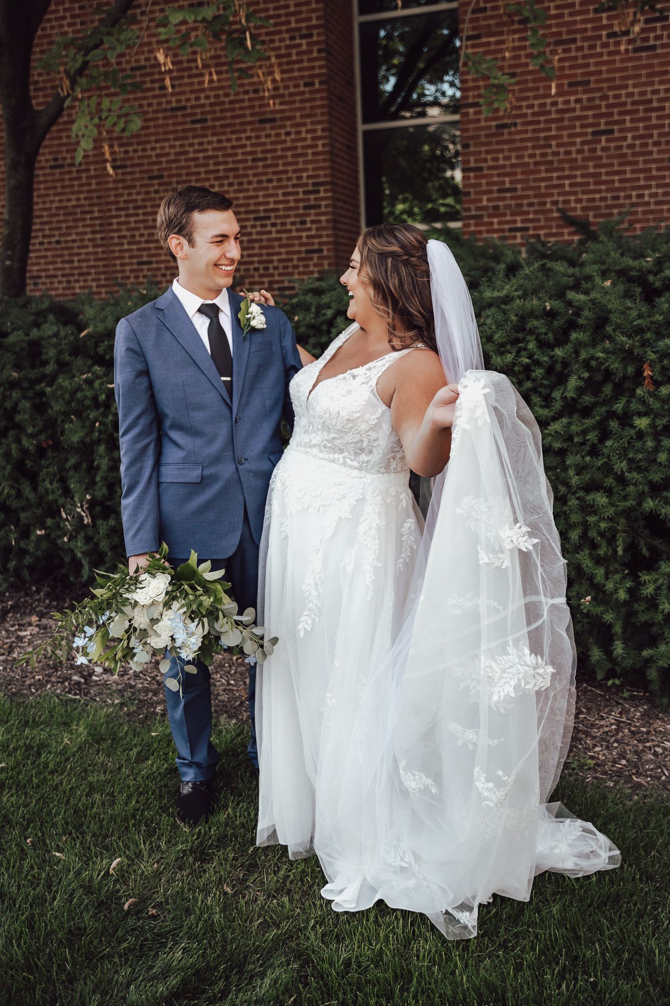 Newlyweds share a candid moment outdoors against a red brick wall backdrop.