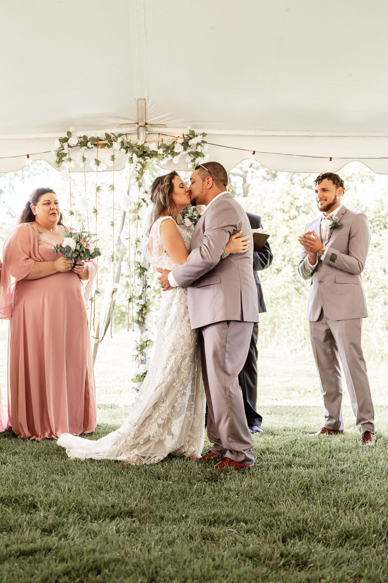 Newlyweds sharing first kiss under tent decorated with greenery and string lights.