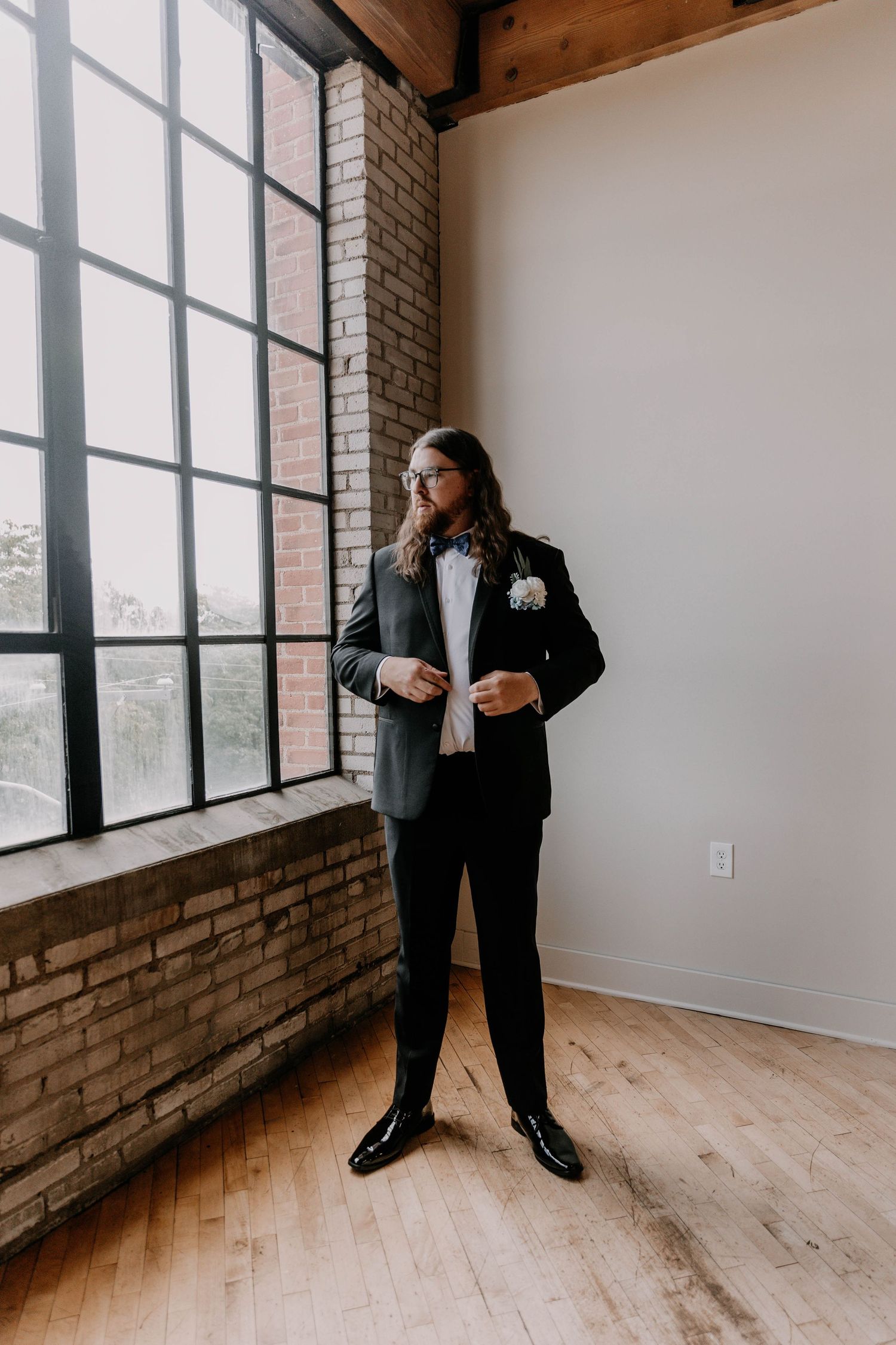 Someone in a classic black tuxedo poses by large industrial windows in a rustic brick loft space.