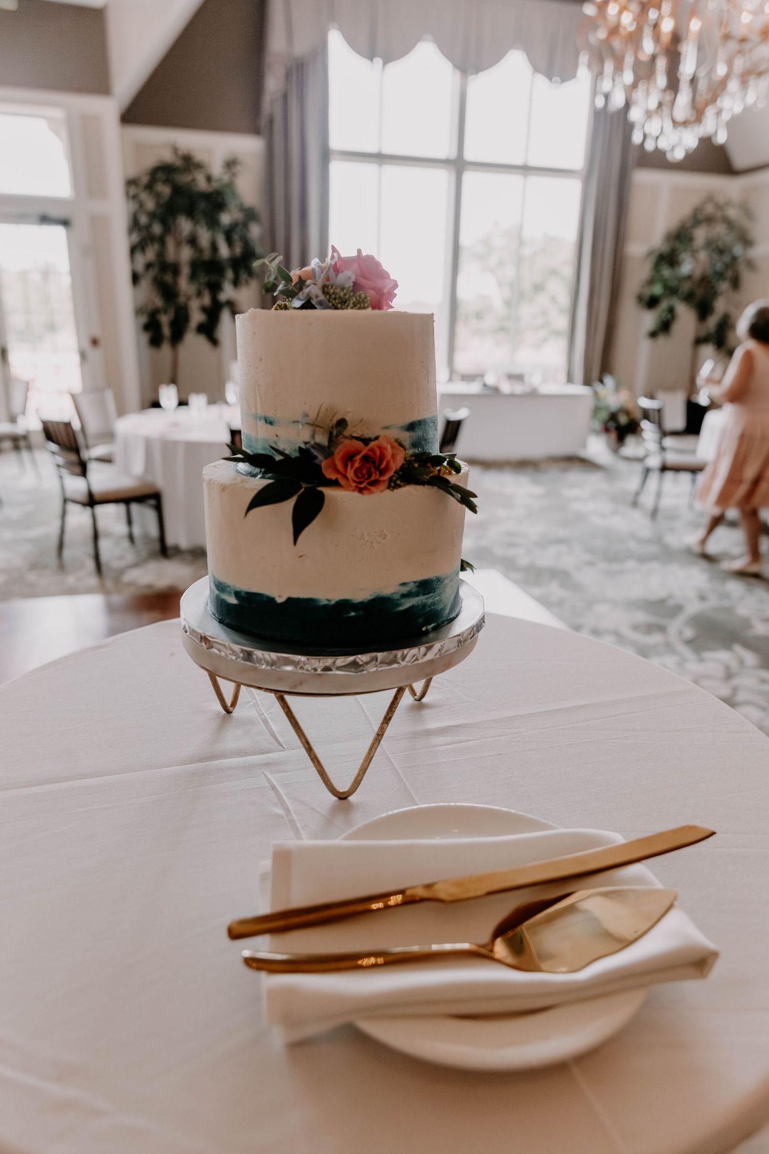 Simple two-tier white wedding cake decorated with fresh flowers on gold stand against bright window.