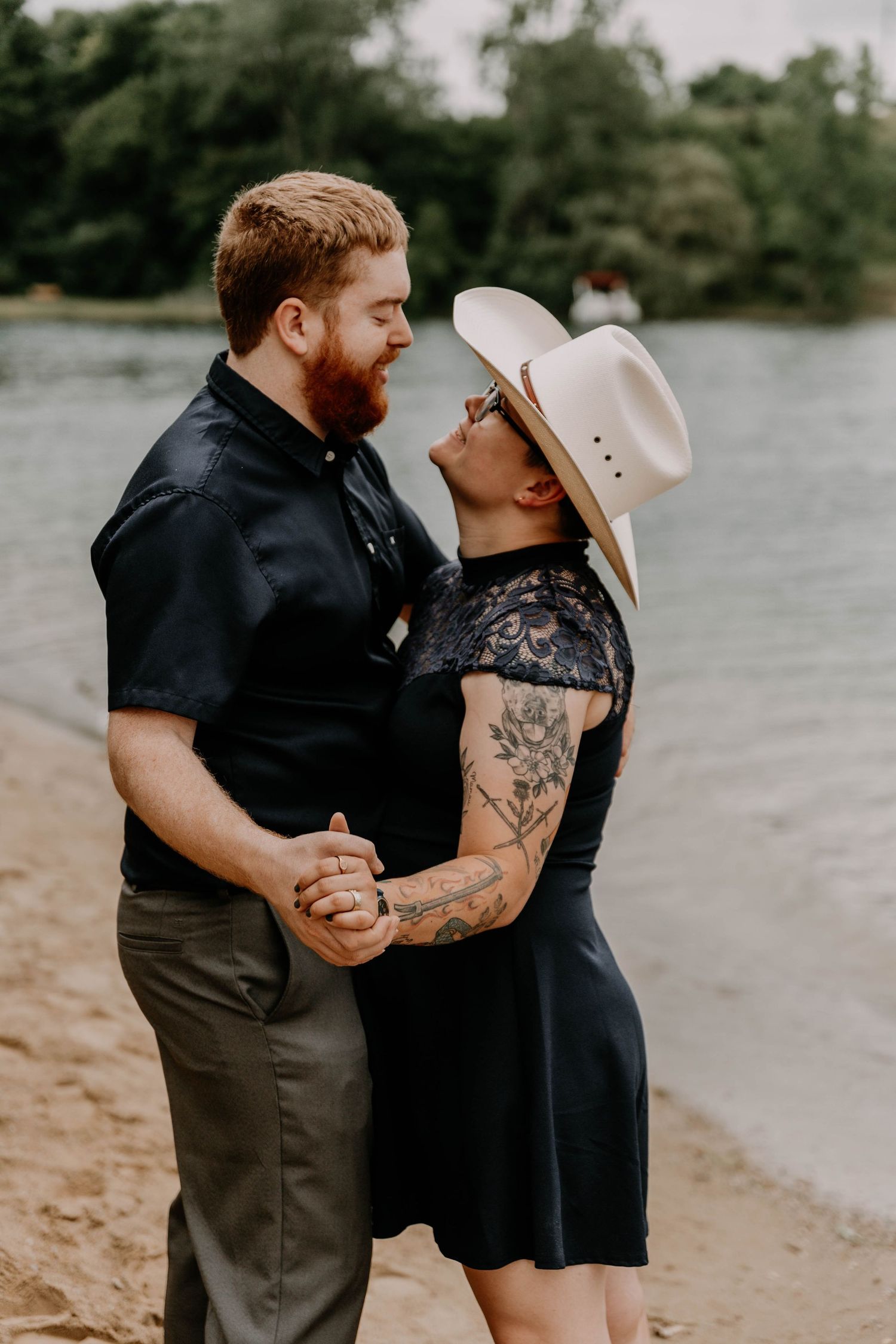 Couple embraces on beach with lake and forest in background, displaying affectionate moment.