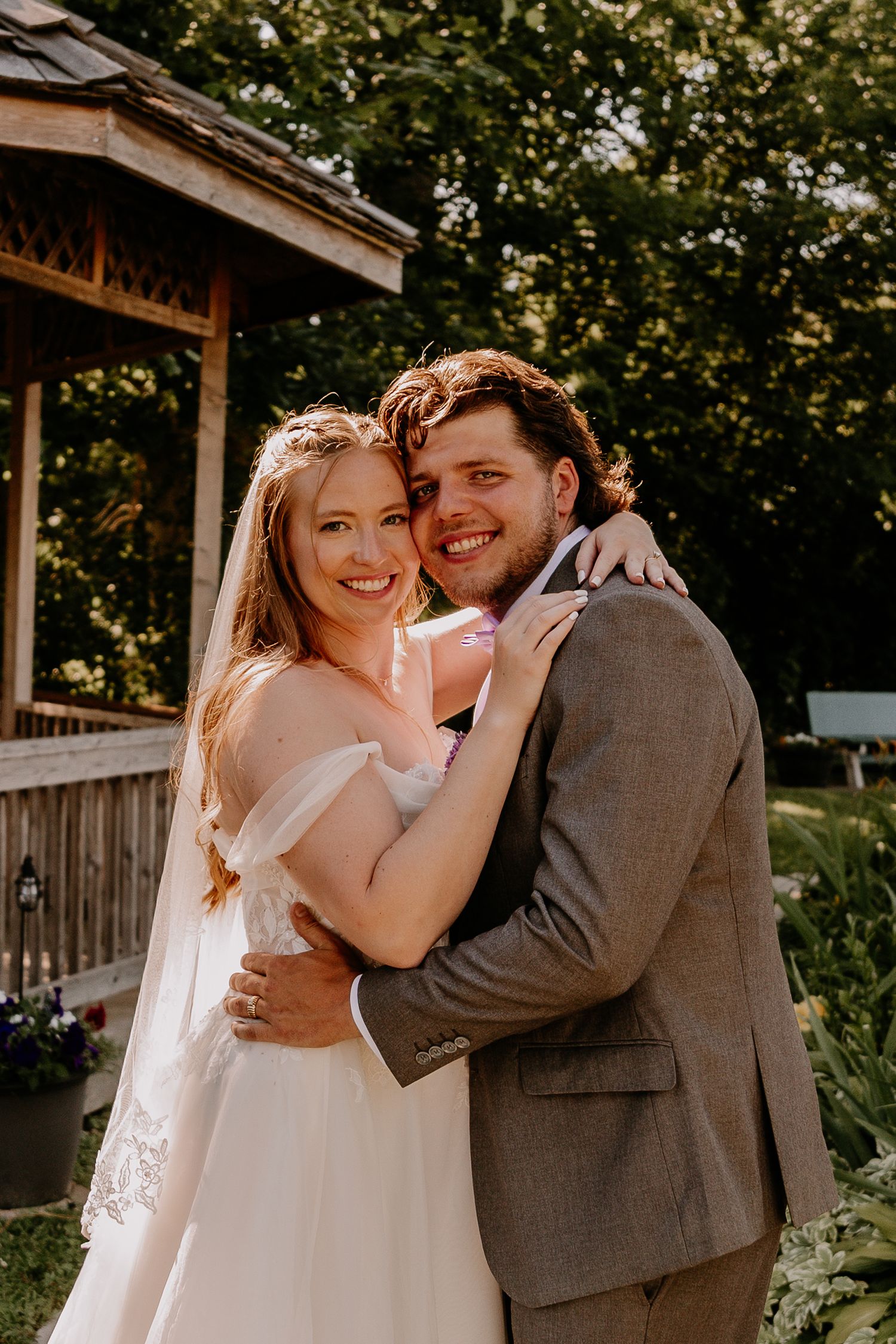 A smiling bride and groom embrace outdoors near a wooden gazebo on their wedding day.