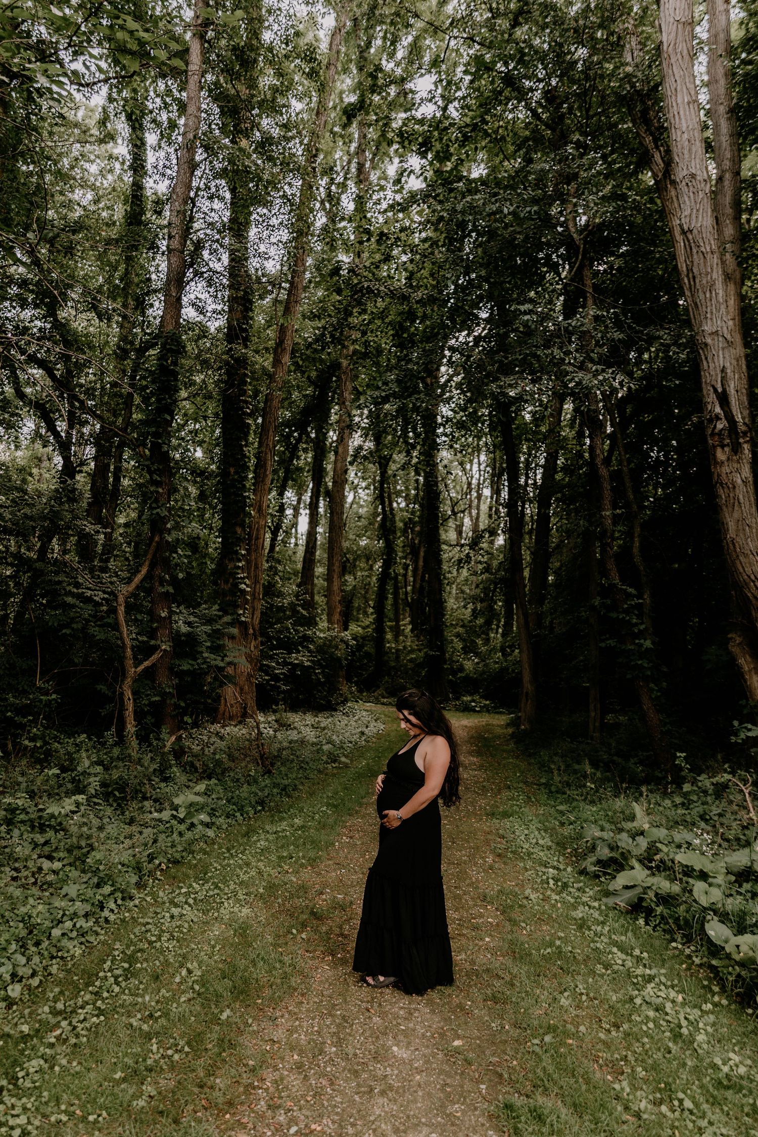 Pregnant woman in black dress standing on forest path surrounded by tall green trees.