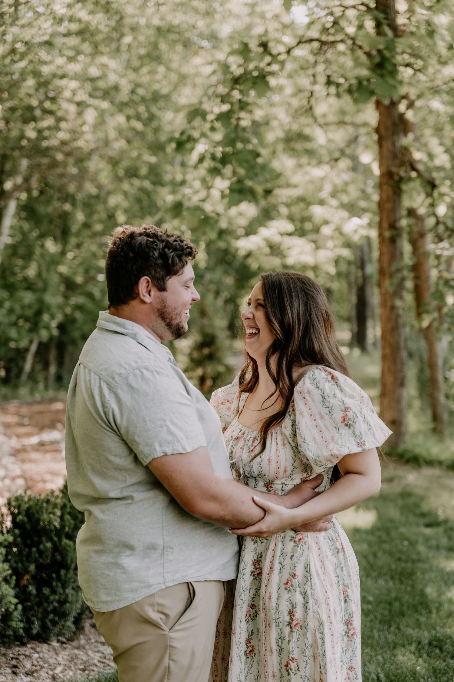 A joyful couple laughs together while embracing outdoors among lush green trees in natural light.