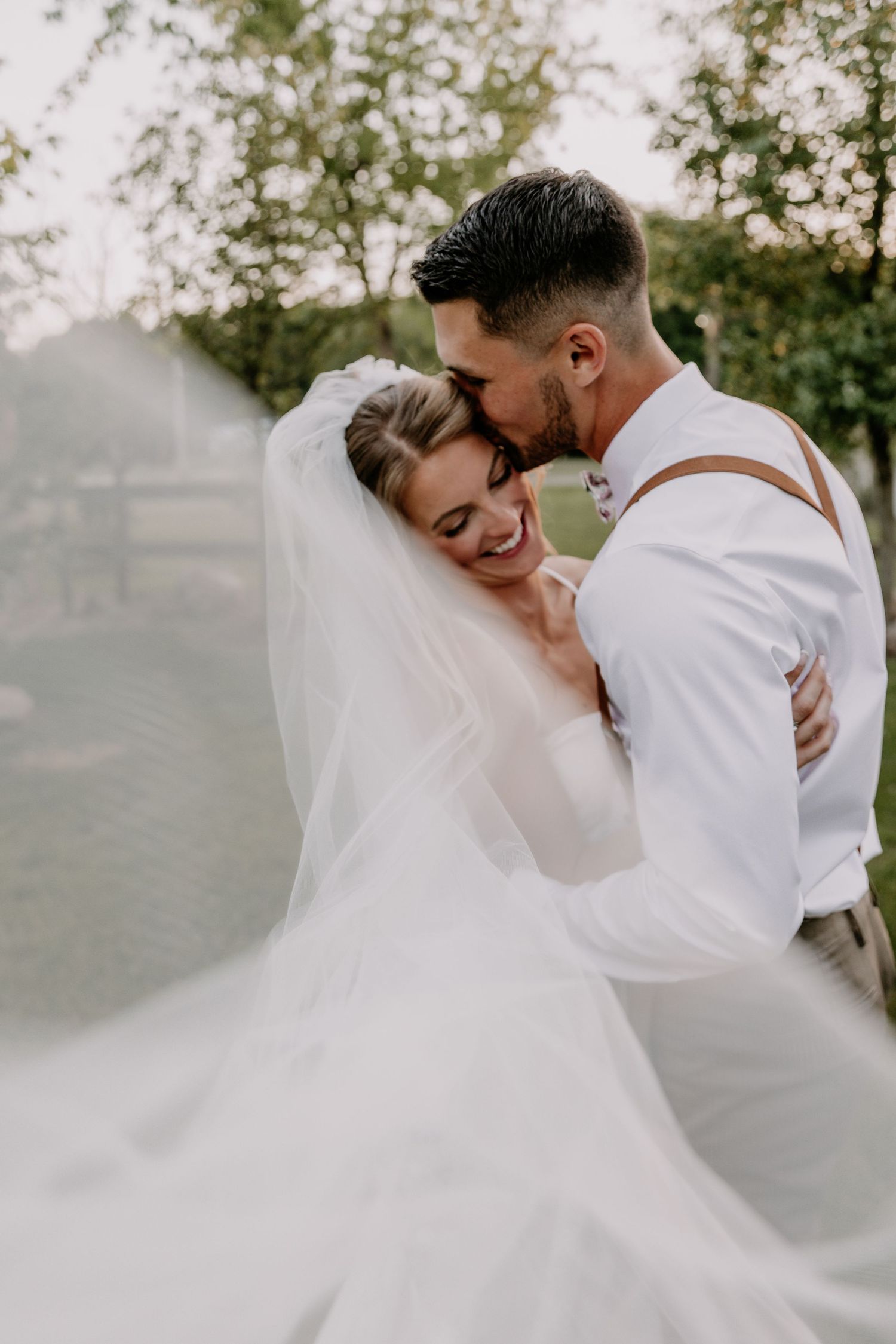 Groom kisses smiling bride on temple as flowing veil billows around them in soft outdoor wedding portrait.