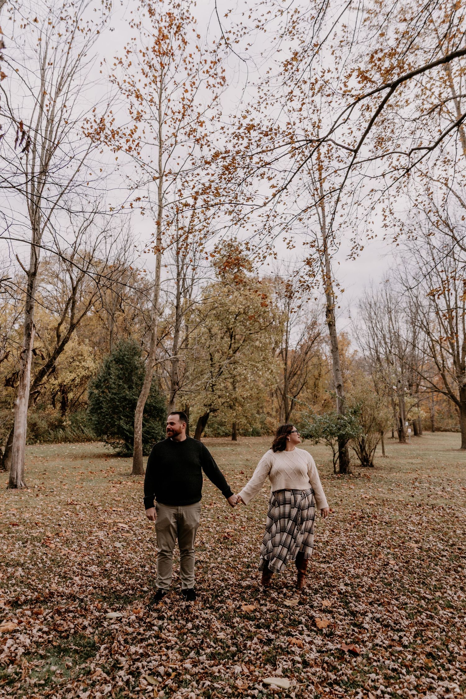A couple walks together through a wooded park covered in fallen autumn leaves on an overcast day.