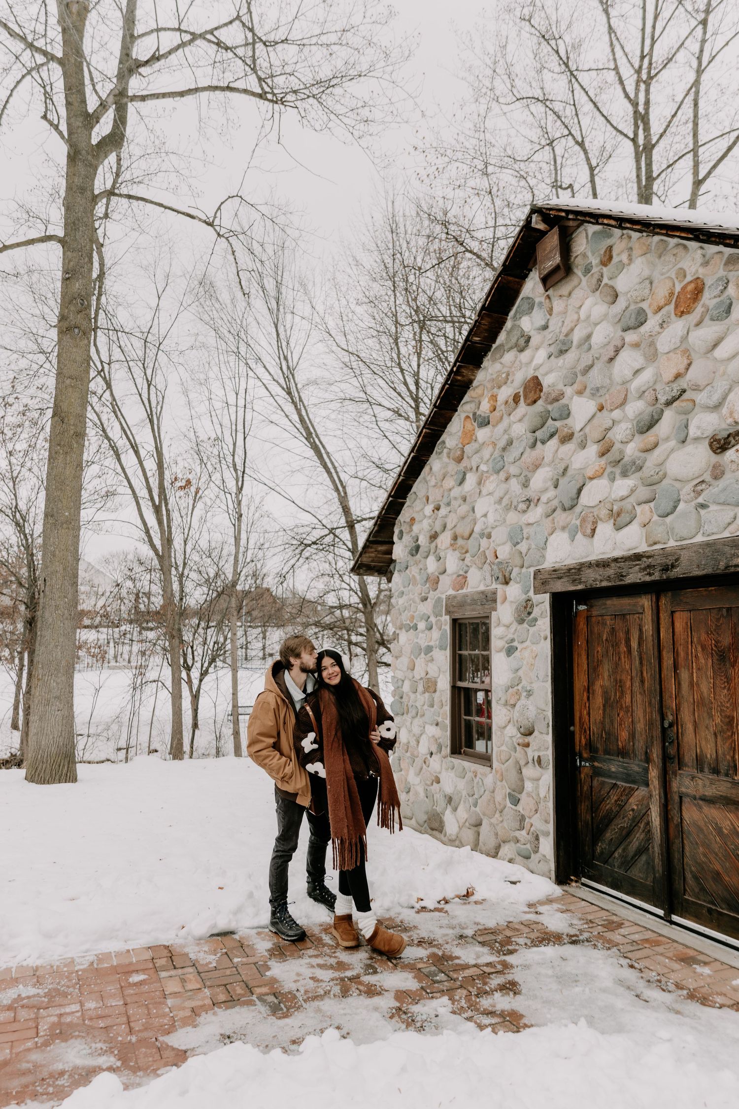 A couple shares a romantic winter moment in front of a rustic stone cabin surrounded by snowy trees.