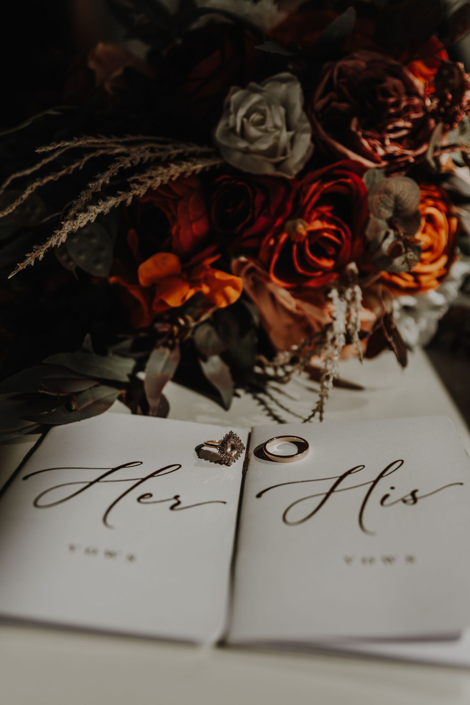 Wedding rings rest on elegant His and Her cards surrounded by moody dark floral decorations in a romantic setting.