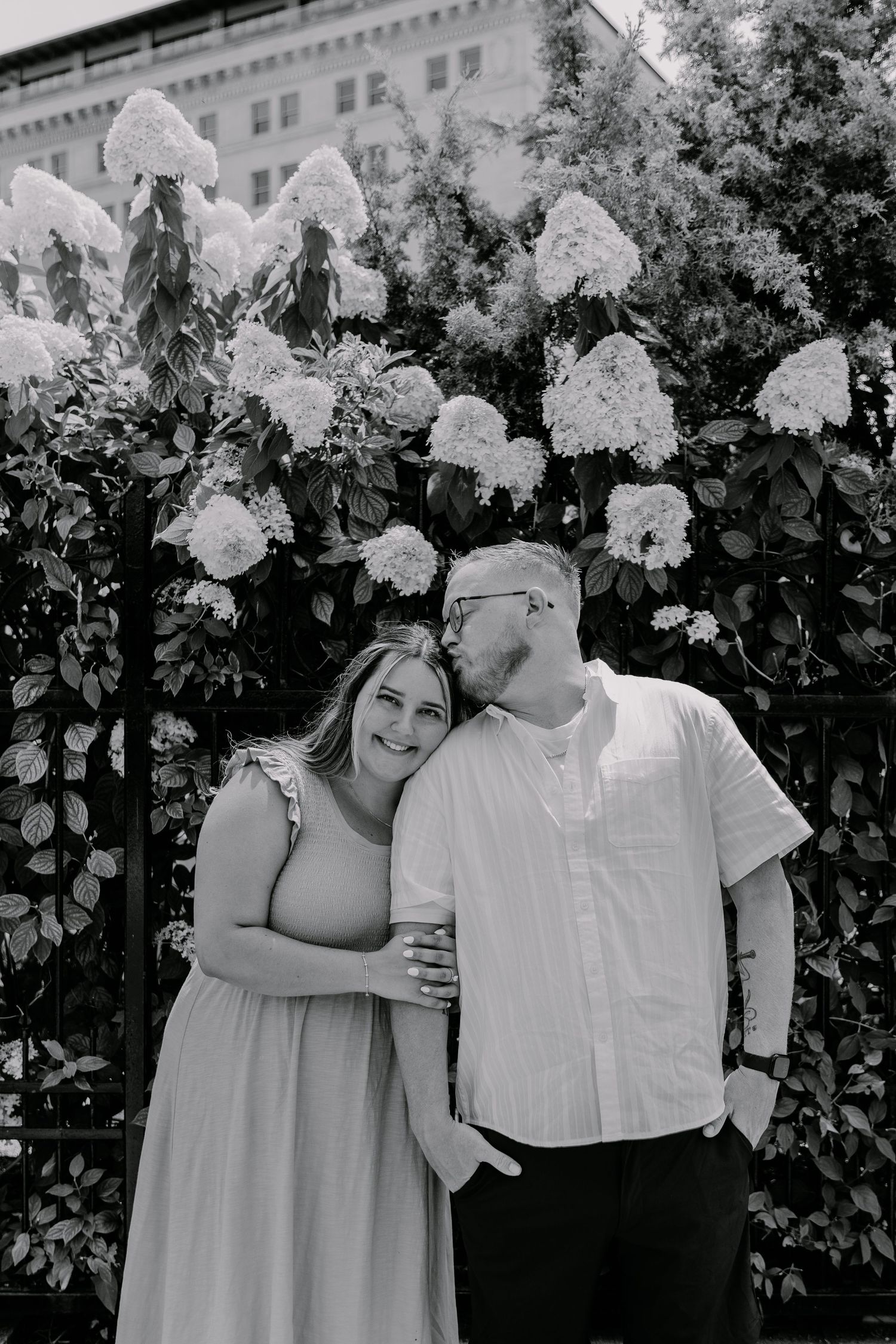 A couple embraces in front of a blooming hydrangea bush with white flowers in a black and white photograph.