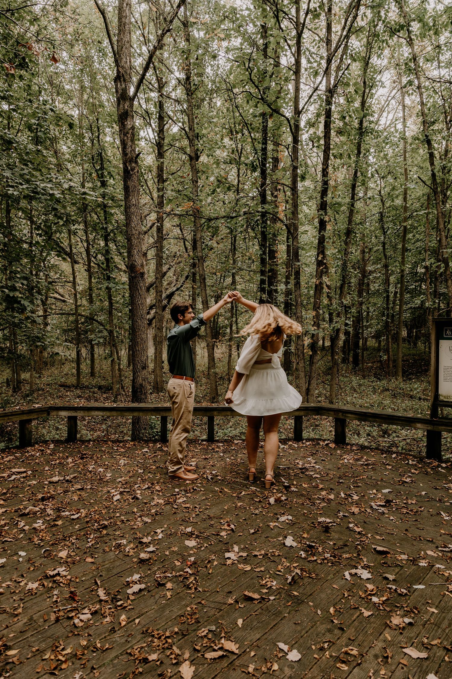 Couple dancing playfully on wooden platform surrounded by trees during photoshoot.