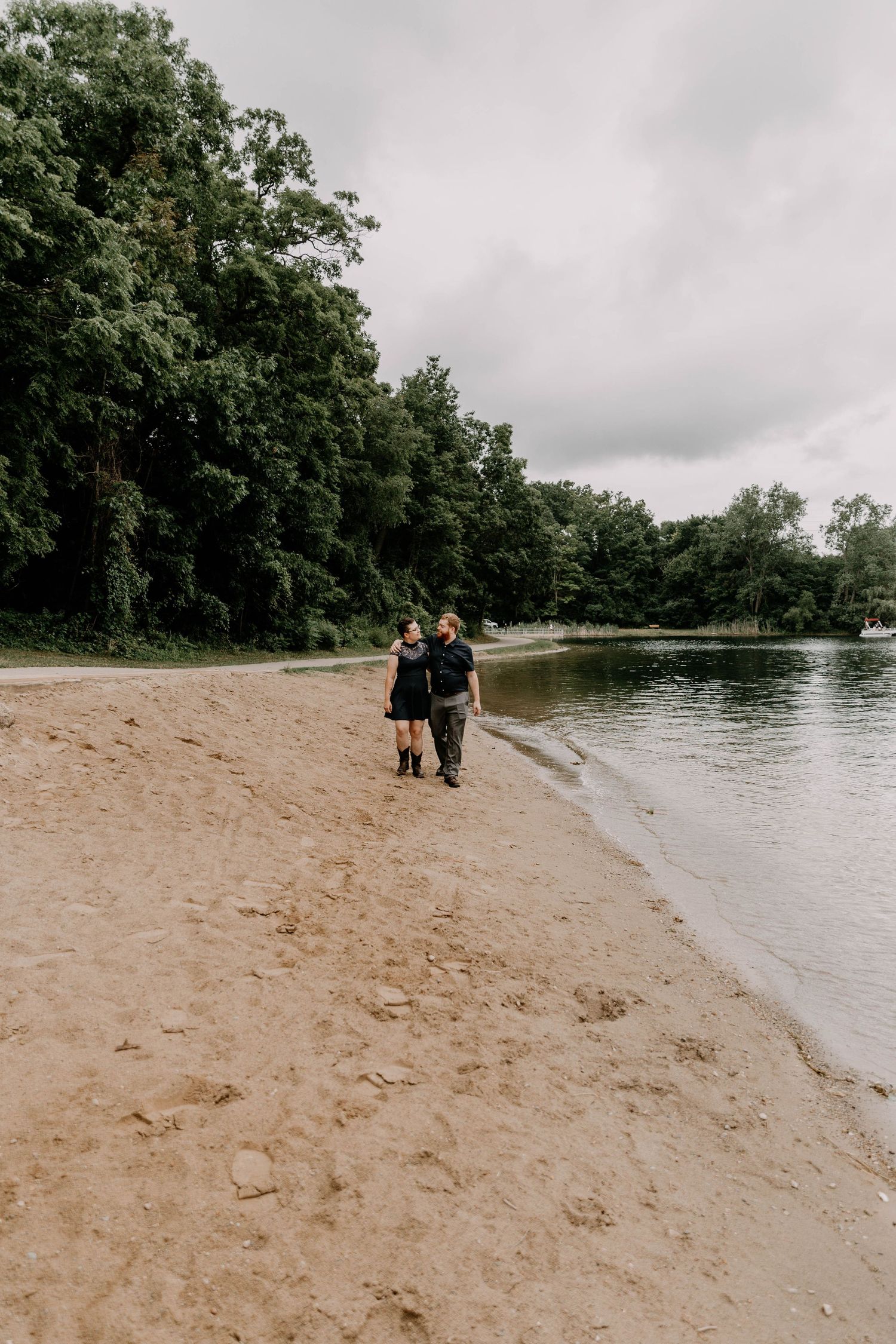 Two people walk together along a sandy beach beside a peaceful lake.