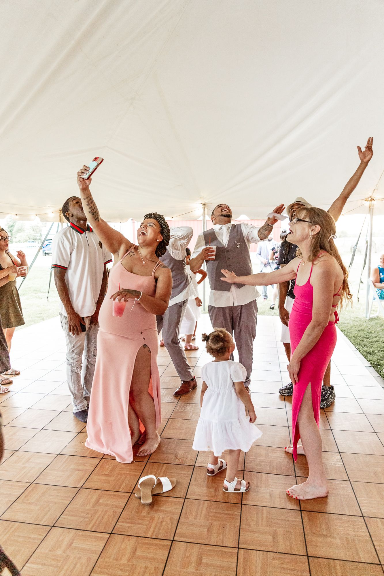 Wedding reception guests dancing together energetically on wooden dance floor under white tent.