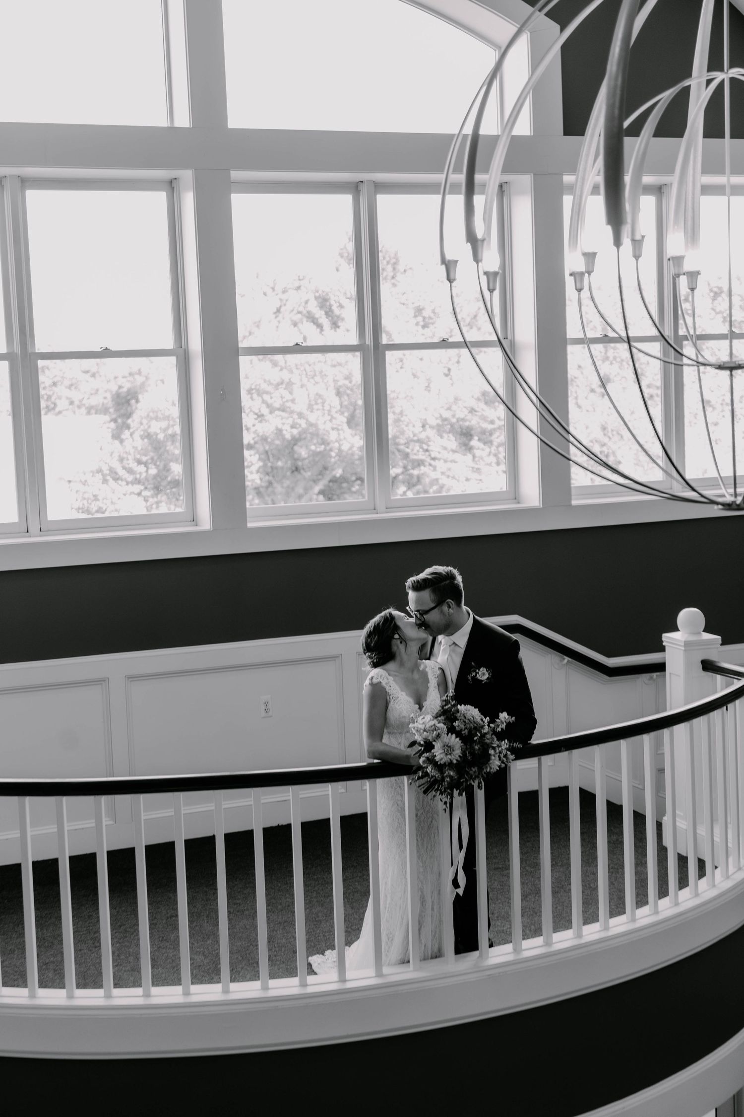 Romantic black and white photo of couple sharing intimate moment on curved white staircase under chandelier.