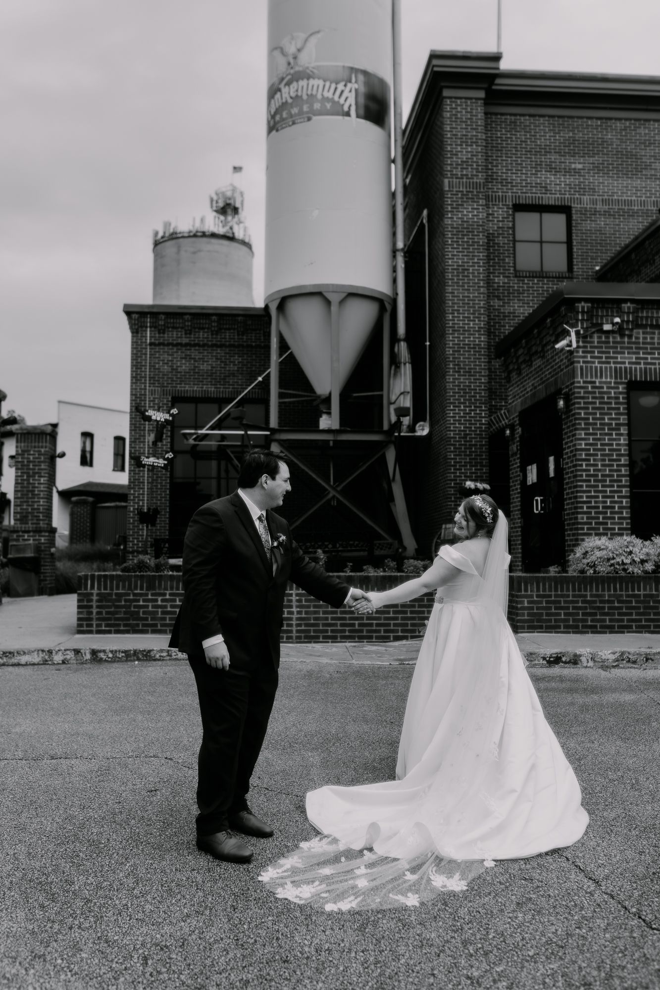 Black and white wedding photo of couple holding hands in front of industrial silos and brick building.