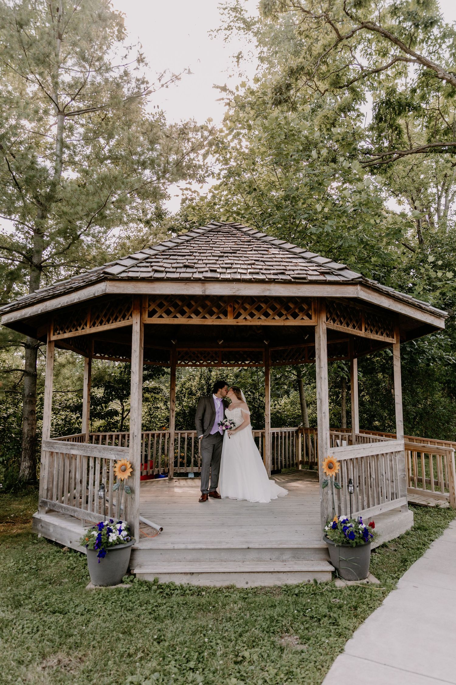 Bride and groom share a kiss in a wooden gazebo decorated with sunflowers and blue flowers surrounded by lush green trees.