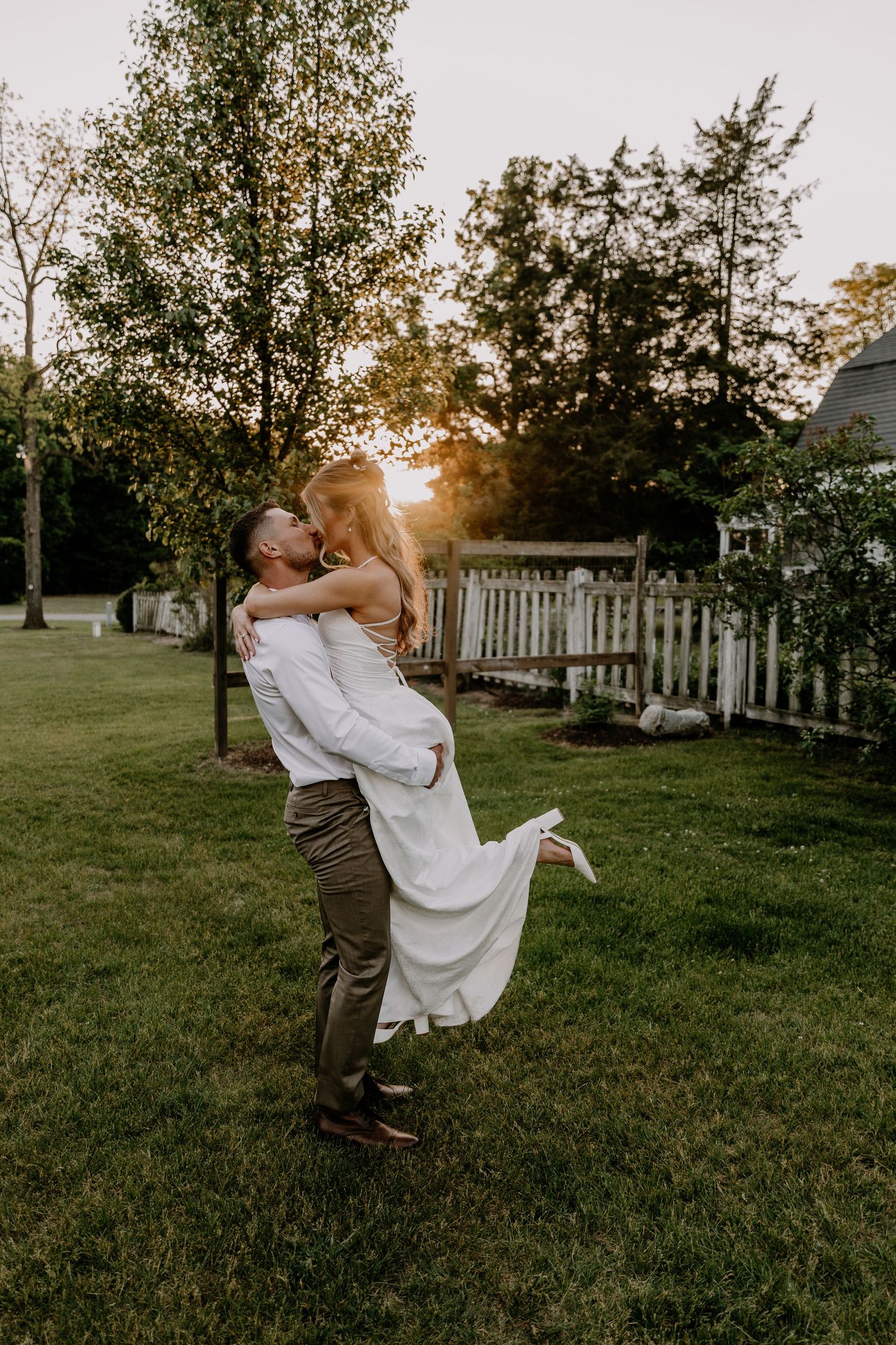 Couple sharing romantic kiss as groom lifts bride in white wedding dress at sunset outdoors.