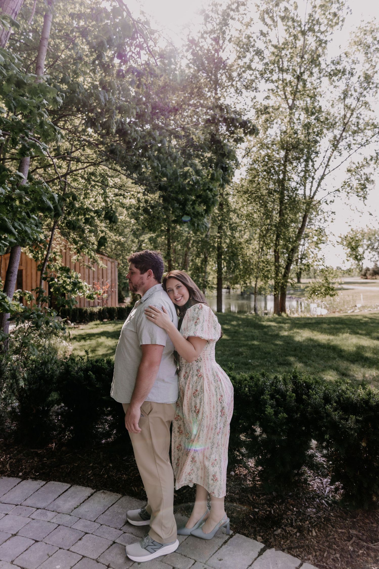 A couple embraces on a brick path in a sunlit garden, the woman wearing a floral dress.