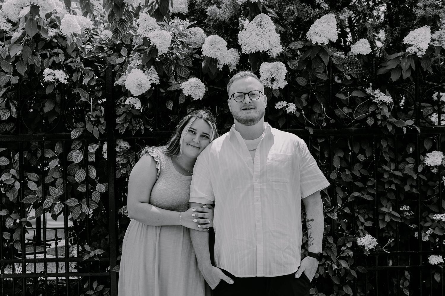 A black and white photo series of a stylish couple posing outdoors in a garden setting with lush foliage.