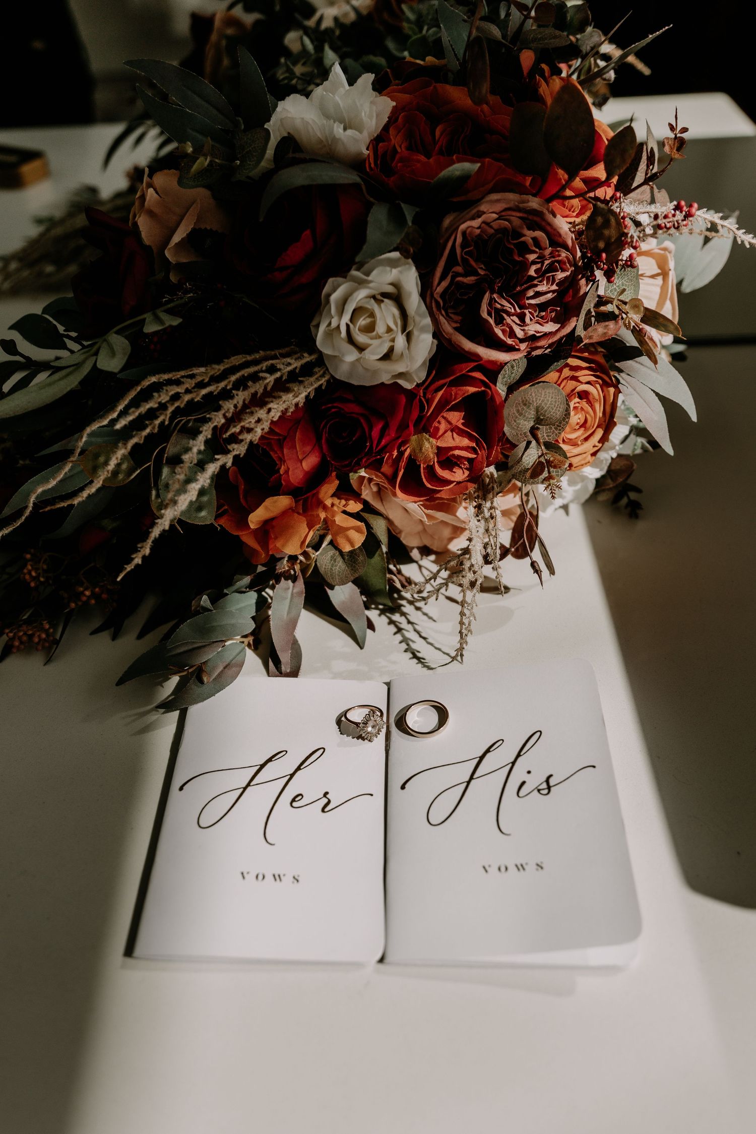 Wedding vow books with elegant calligraphy lie beneath a romantic arrangement of red and white roses and greenery.