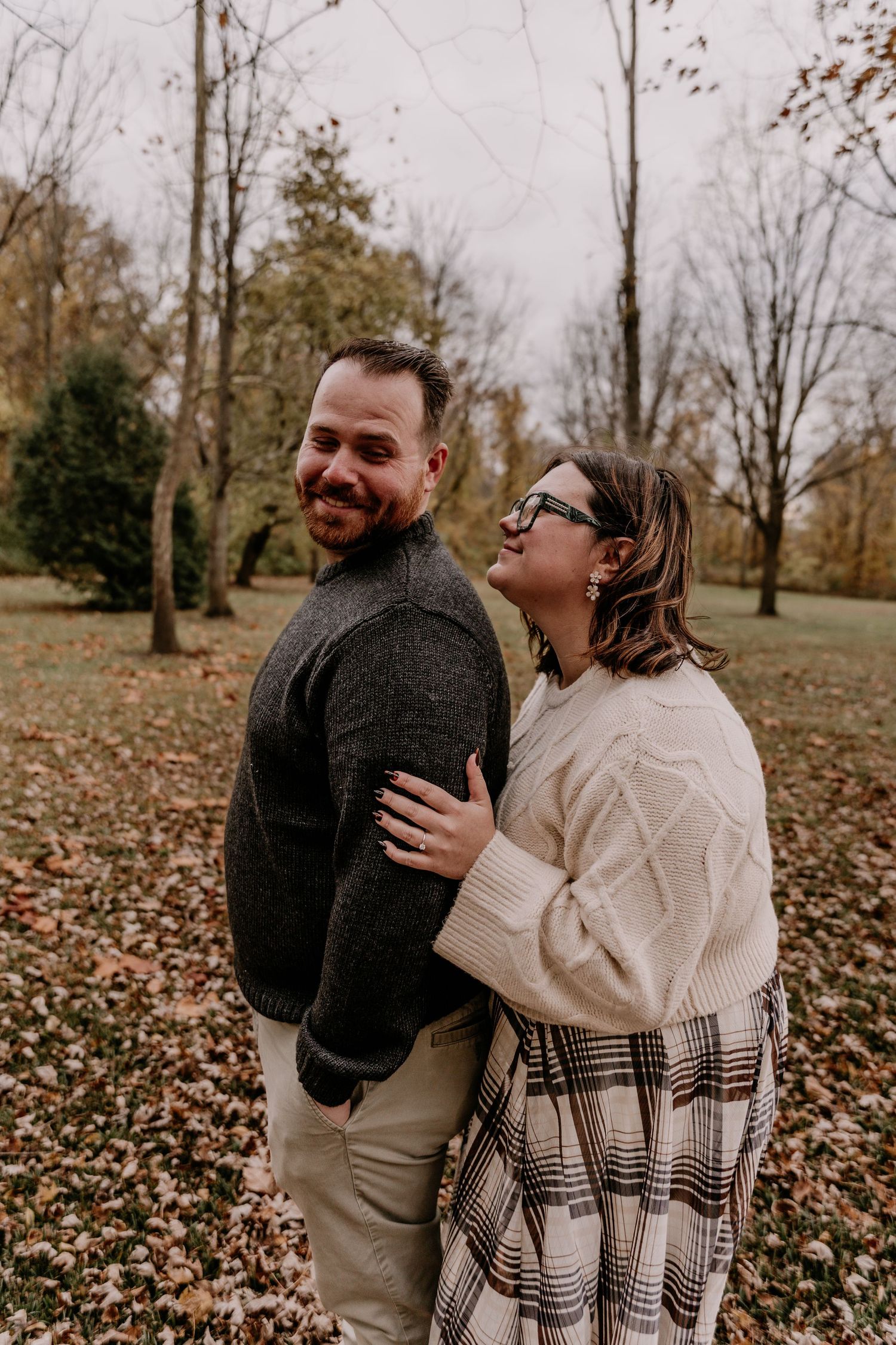 A couple shares a joyful moment together in a park filled with autumn leaves on a cloudy day.