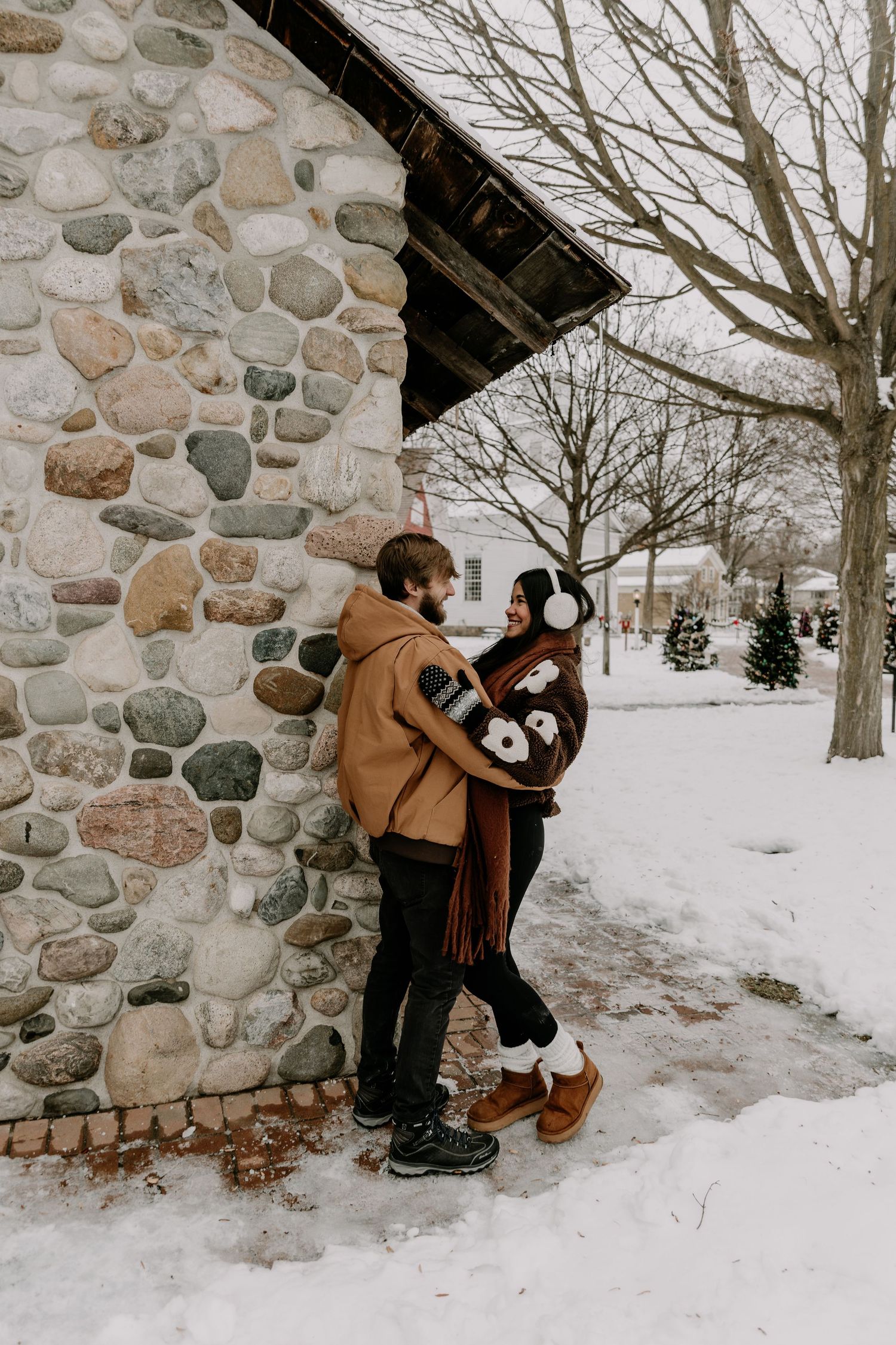 A romantic winter engagement photo series with a couple embracing against a rustic stone wall in the snow.
