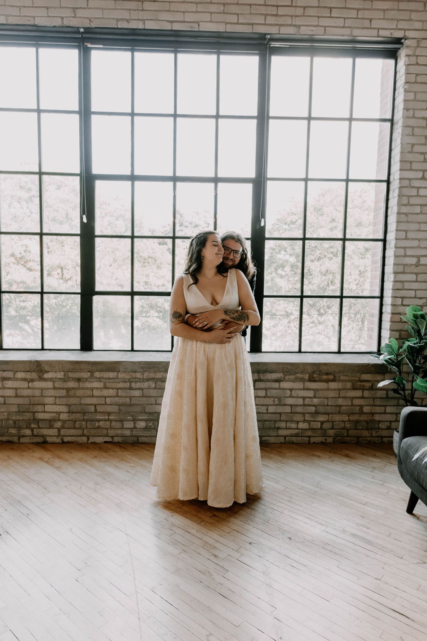 A couple embraces in an elegant wedding venue with large windows and exposed brick.
