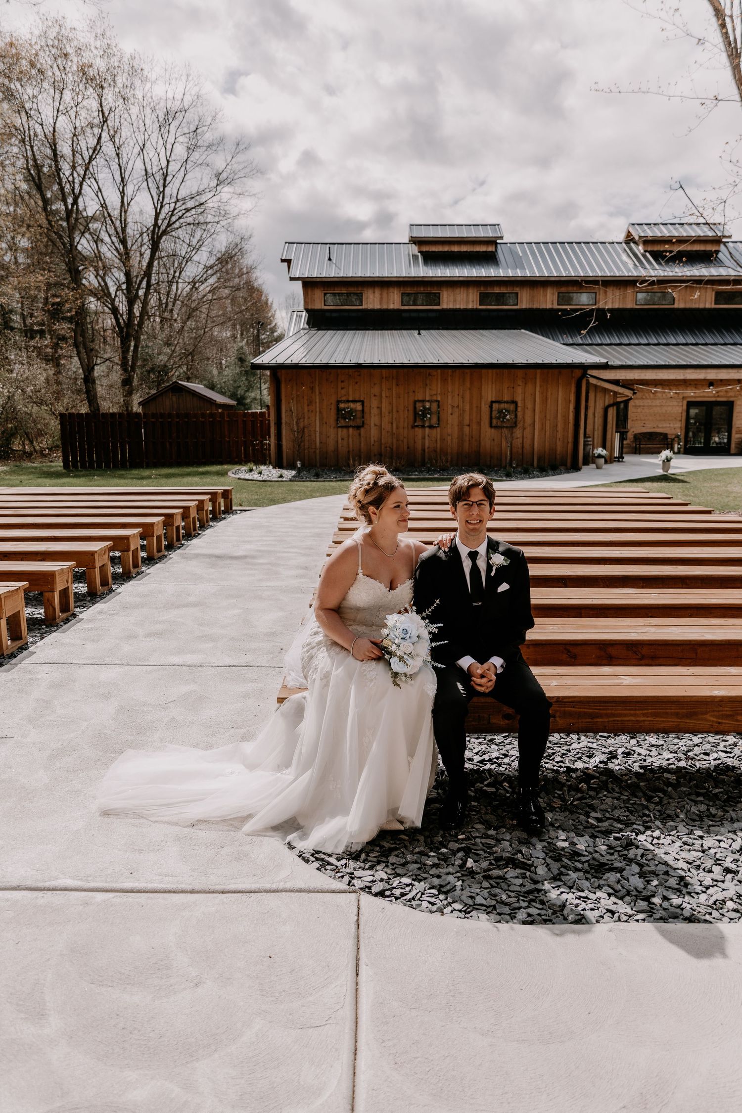 Wedding couple seated at outdoor ceremony venue with wooden benches and modern architecture.