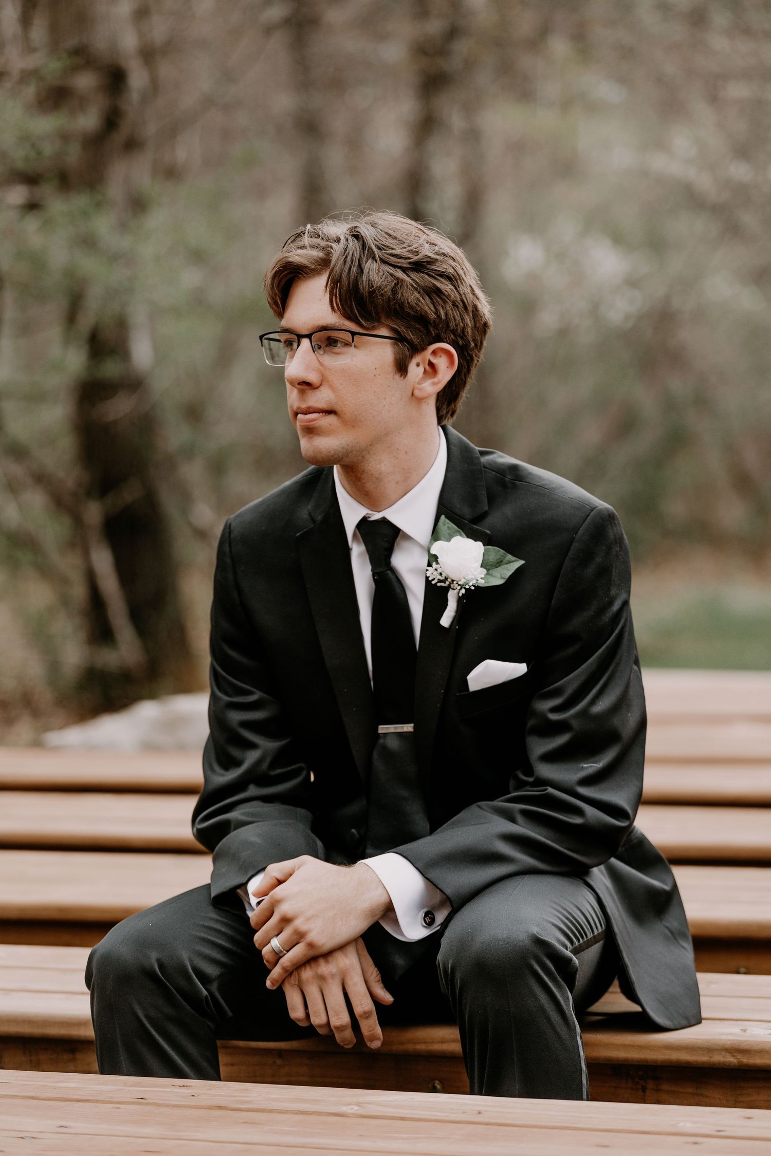 Wedding party member in black suit sitting on wooden bench outdoors.
