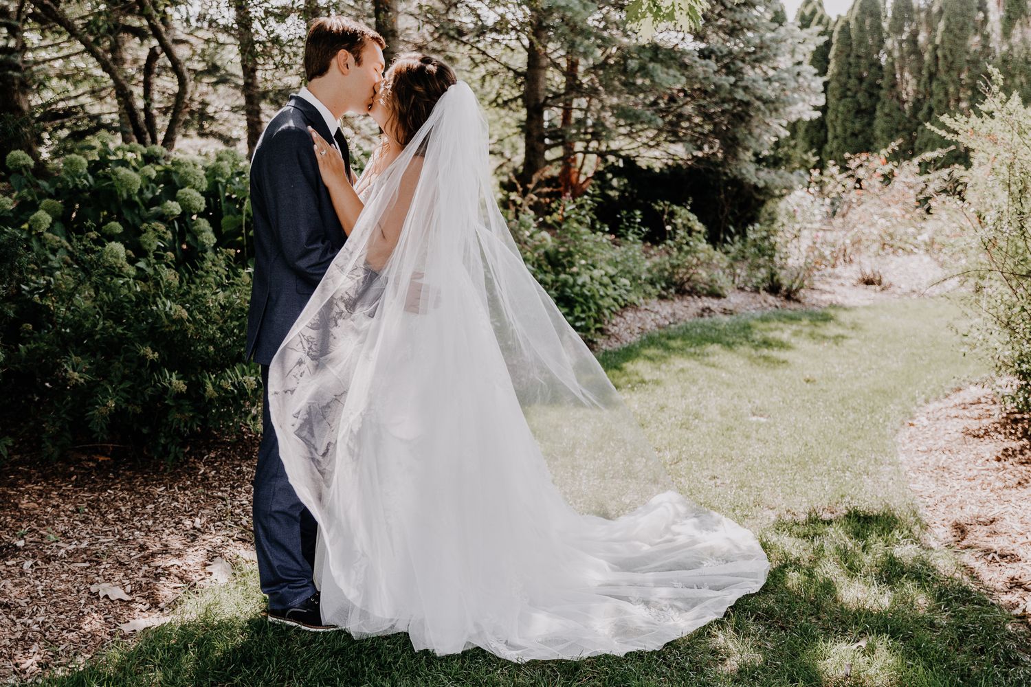 Elegant couple shares romantic kiss in garden setting with flowing white wedding dress and veil.