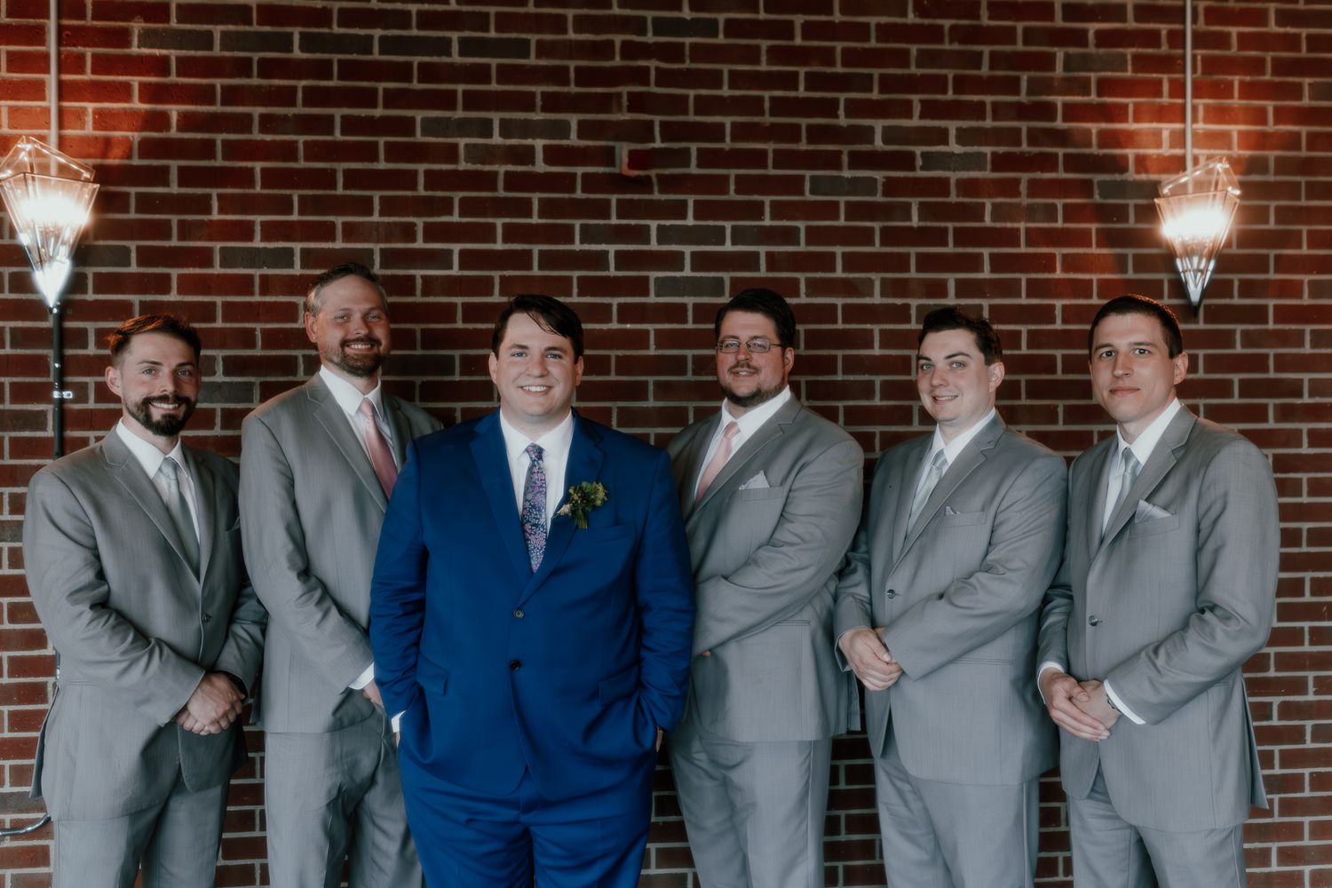 Groomsmen group photo with one in navy suit surrounded by others in light grey suits against brick wall.
