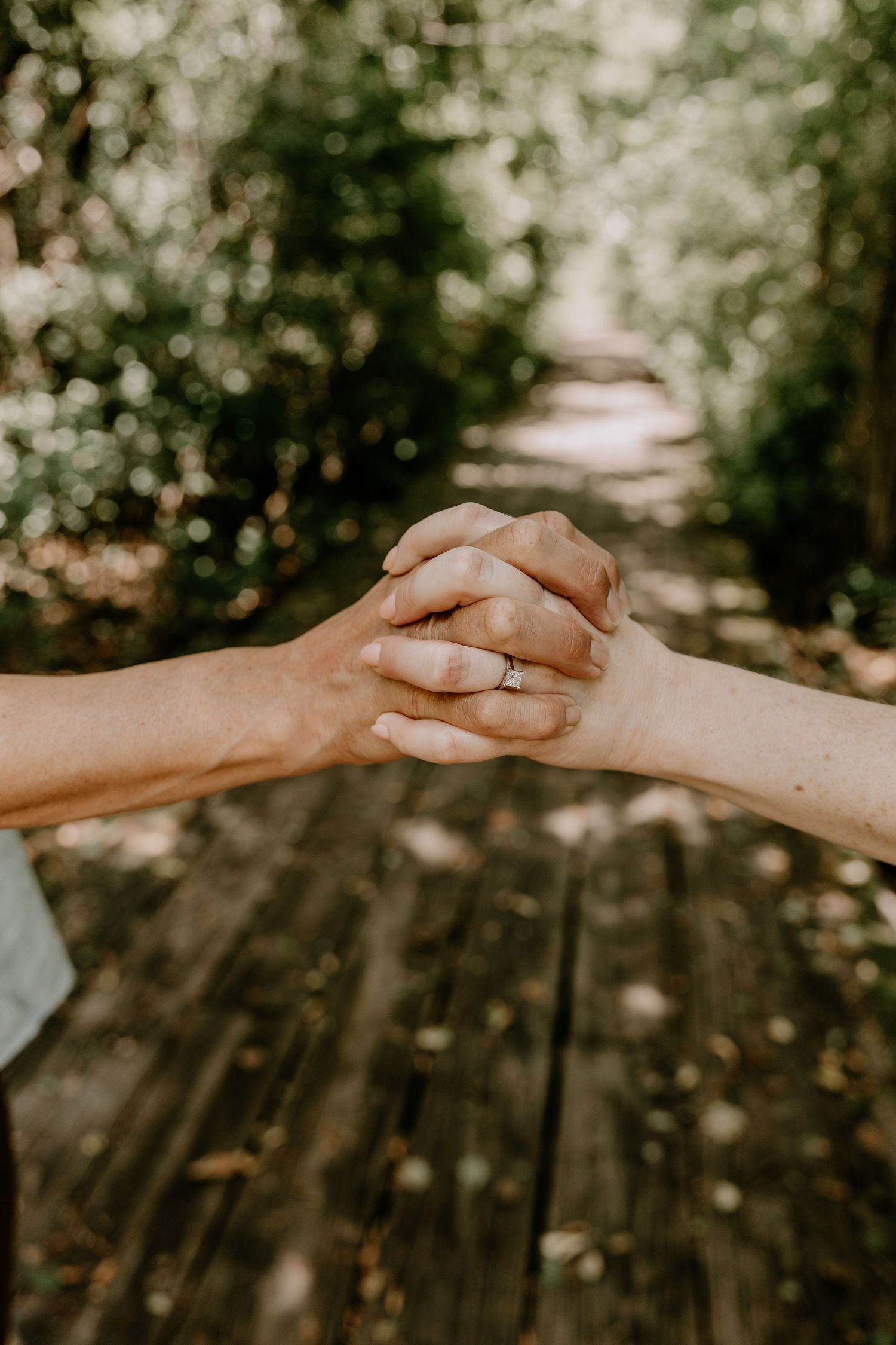Two hands tenderly clasped together against a sunlit wooded background, with one hand wearing an engagement ring.