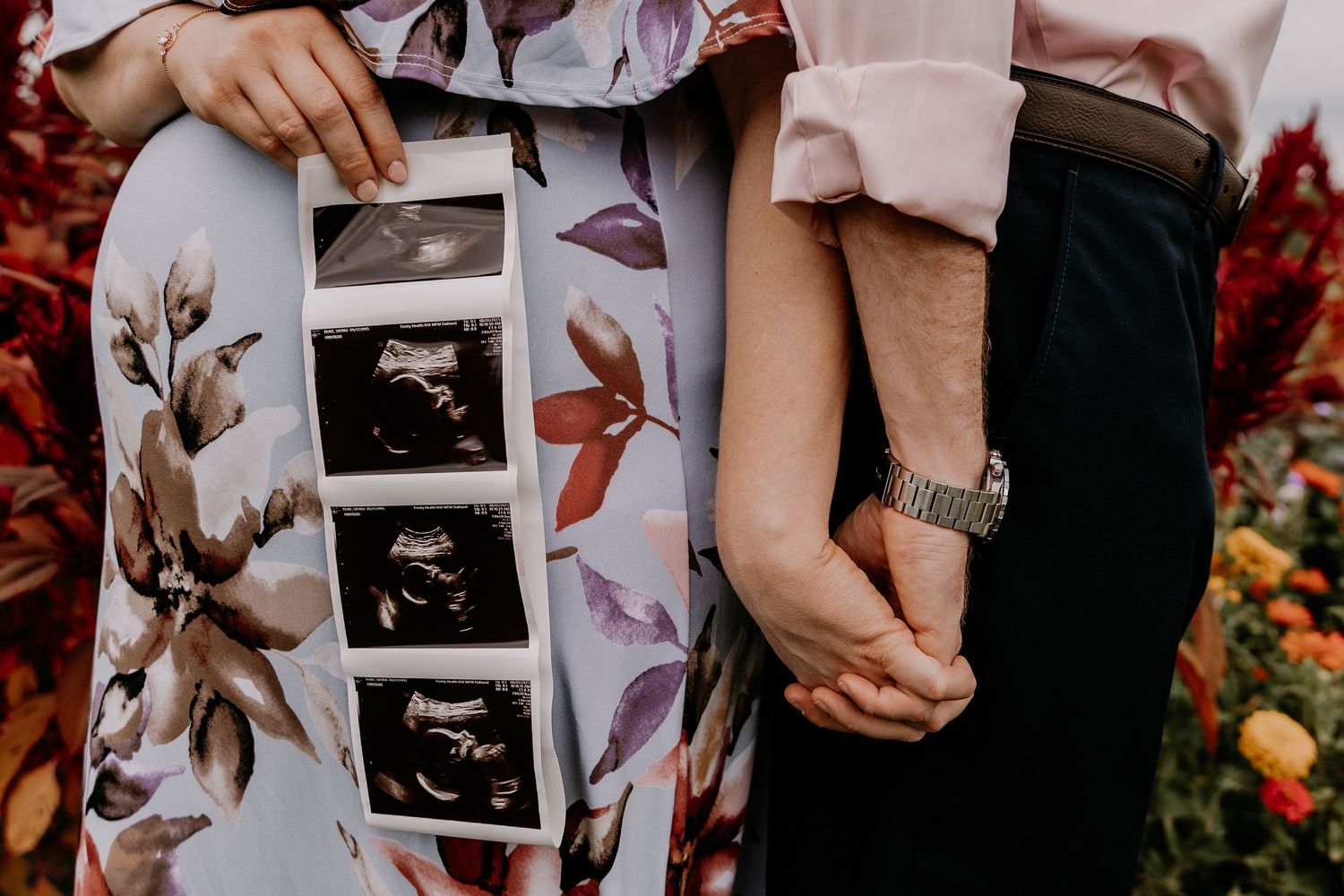 A close-up view of hands holding black and white sonogram ultrasound photos amid colorful fall foliage and floral background.