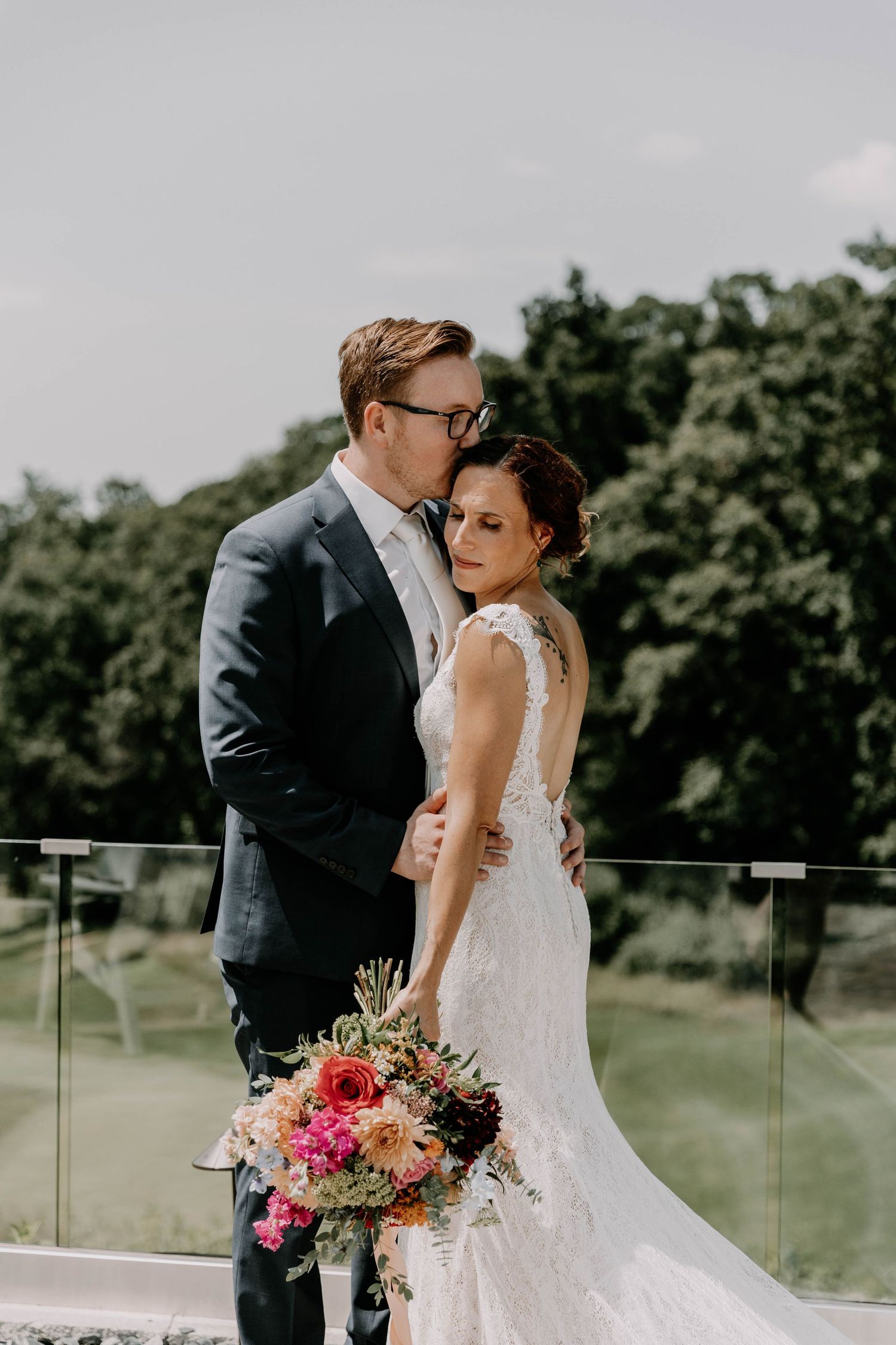 Wedding couple embraces on an outdoor terrace with a colorful floral bouquet featuring pink and red blooms.