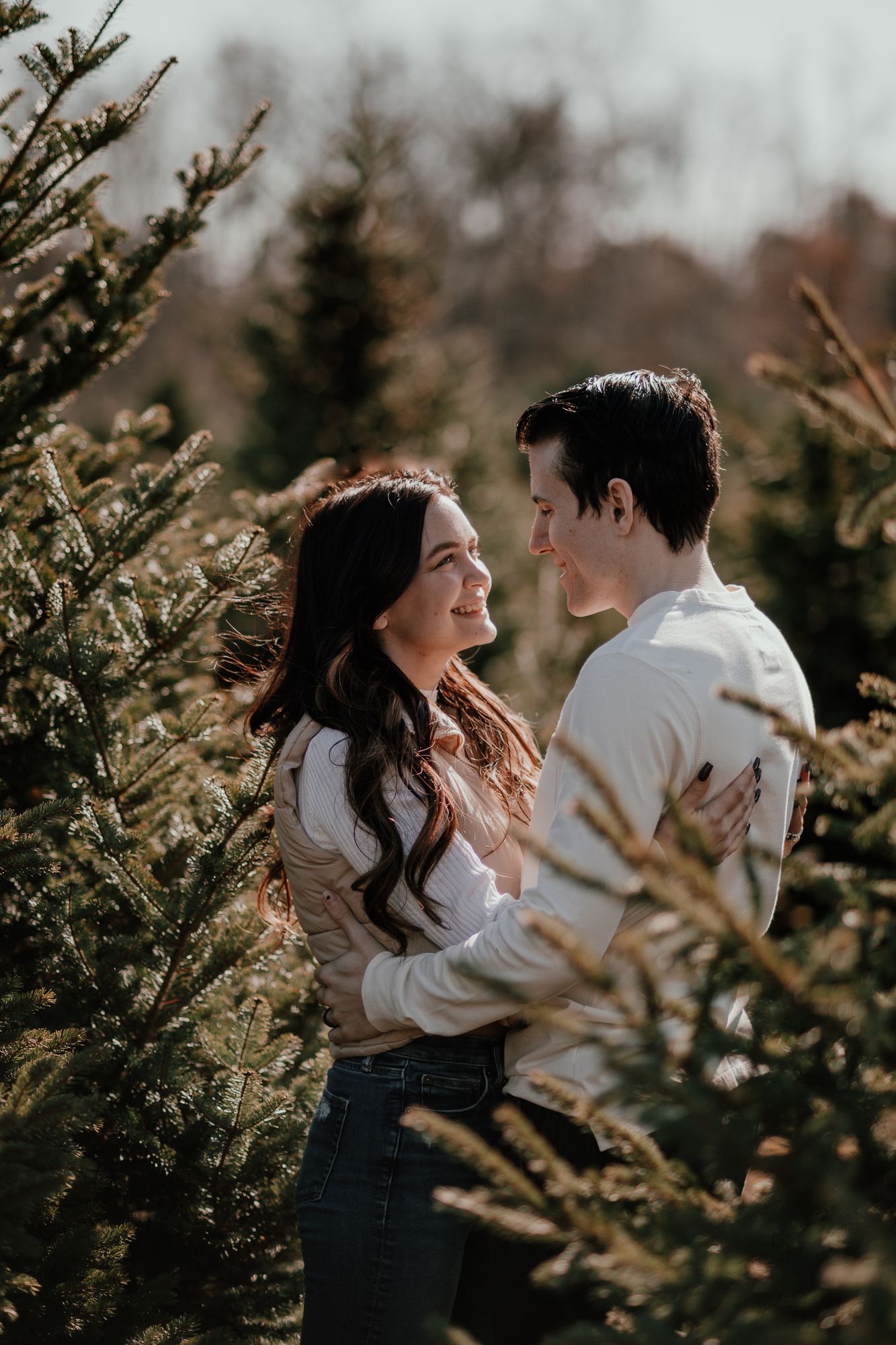 Couple in white sweaters embracing among evergreen trees during romantic photoshoot.