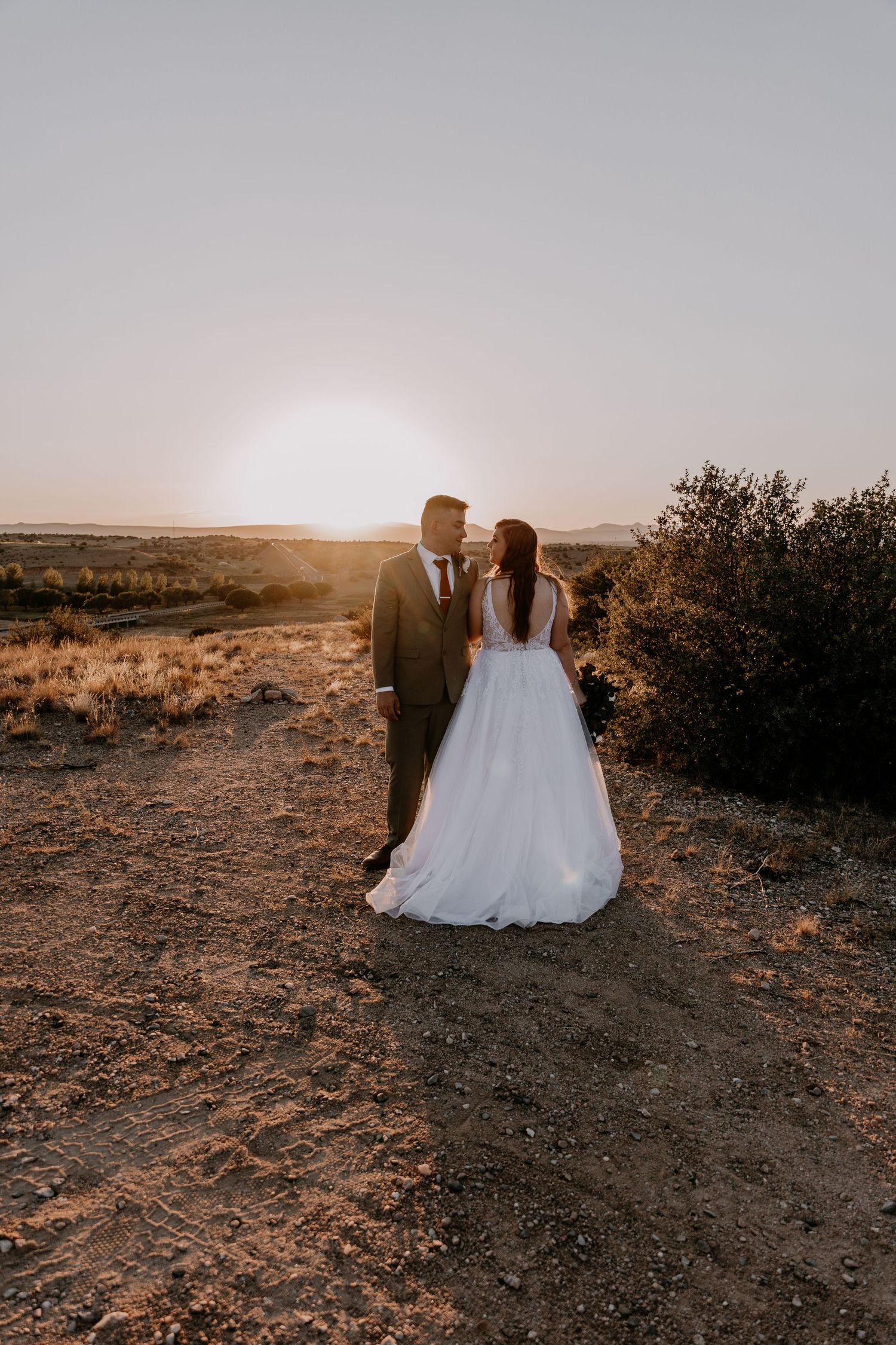 Newlyweds stand together on desert hilltop with golden sunlight behind them.
