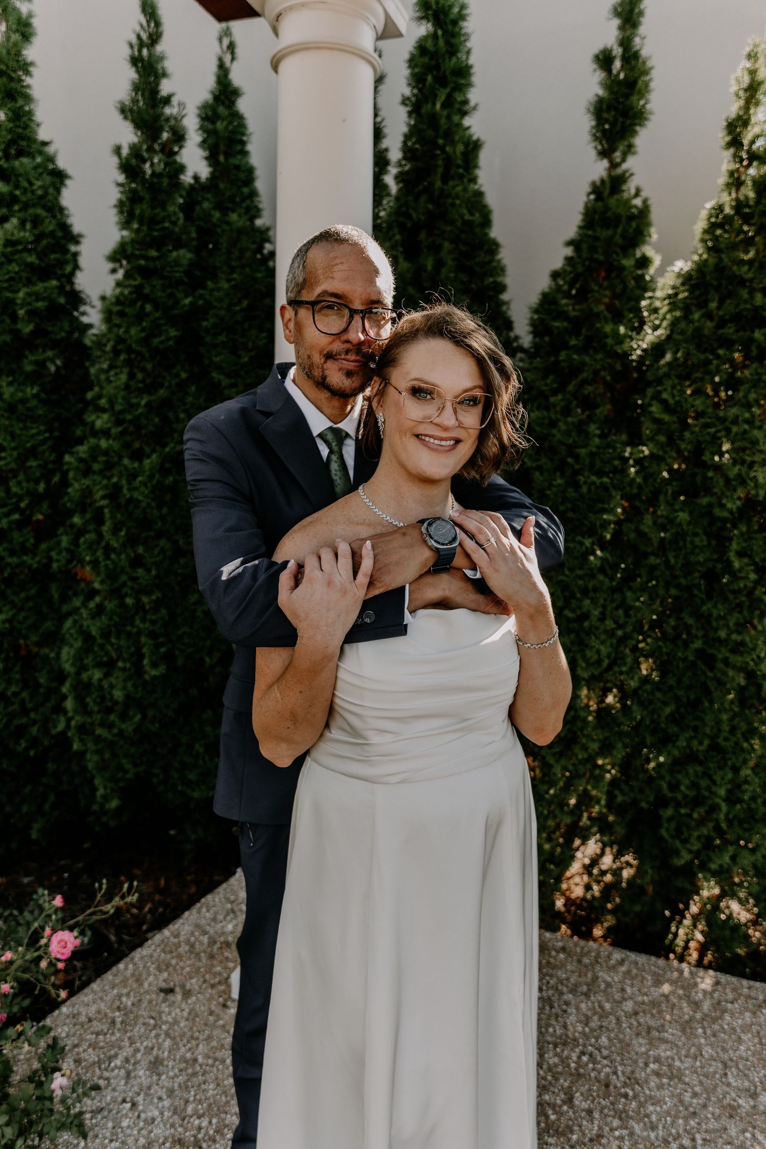 A couple in wedding attire embraces among tall cypress trees and white columns in an outdoor garden setting.