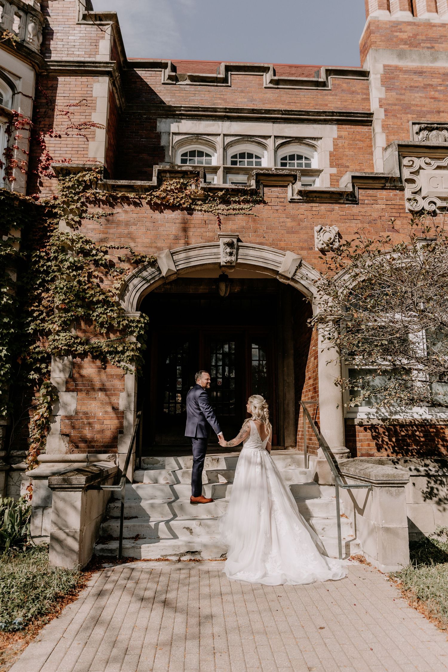Bride and groom hold hands on stone steps of ivy-covered brick mansion wedding venue on sunny autumn day.