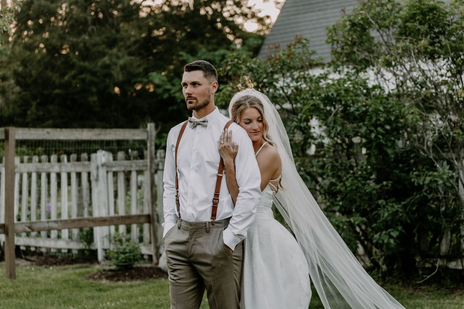 Bride and groom posing outdoors in garden setting, groom in suspenders, bride in white gown with veil.
